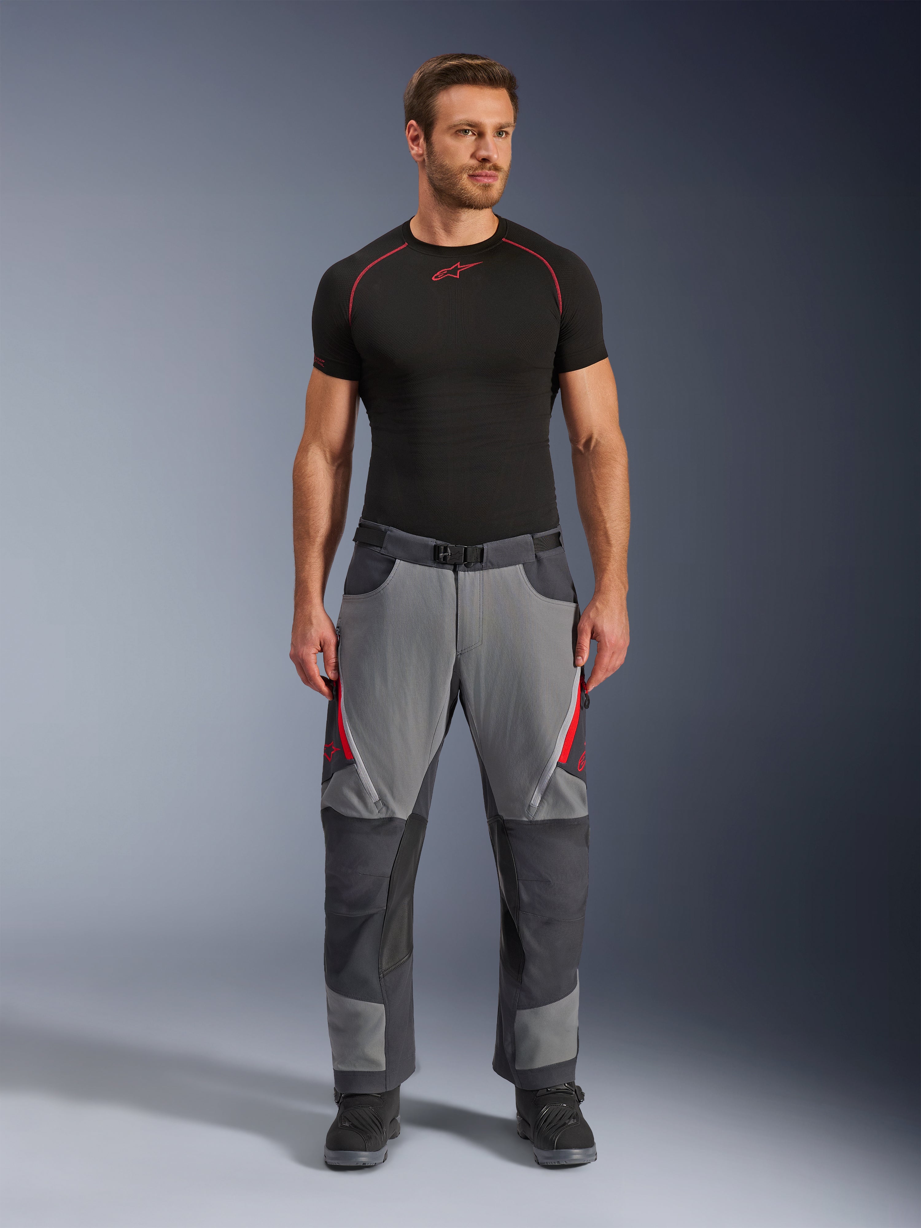 Alpinestars Maxdura Dual bukser, motorcykelbukser, mørkegrå og dyb grå med røde detaljer, set forfra båret af en model med sort Alpinestars-basislag og sorte støvler, med forstærkede paneler og lårventilation med lynlås, adventure-køreudstyr