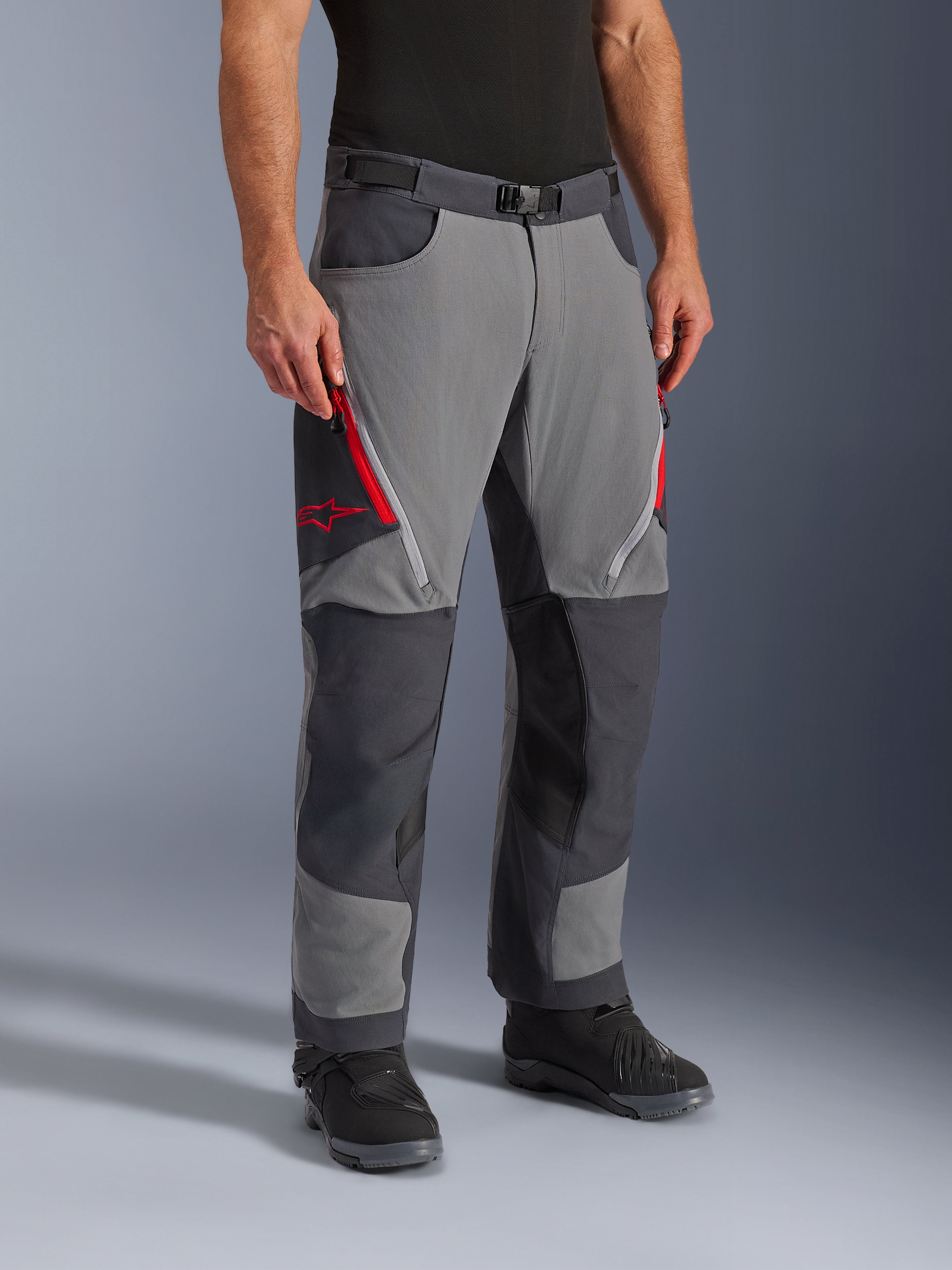Alpinestars Maxdura Dual bukser i mørkegrå og dyb grå, adventure-touring motorcykelbukser med røde lårventiler med lynlås og stjernelogo, forstærkede mørke paneler på knæene, stylet med sorte motorcykelstøvler