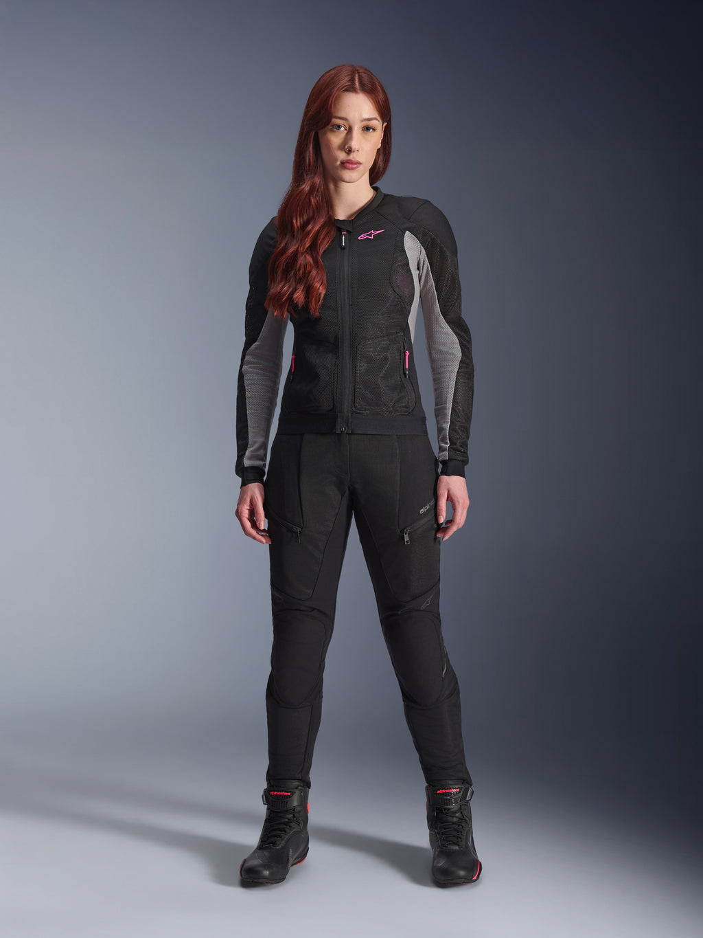 Alpinestars Stella Troop-Air jakke og bukser, motorcykeludstyr til kvinder, sort med Ash Gray og Pink Fluo detaljer, båret af en kvindelig model med rødt hår, med mesh-ventilation og beskyttende design, stående positur i studie