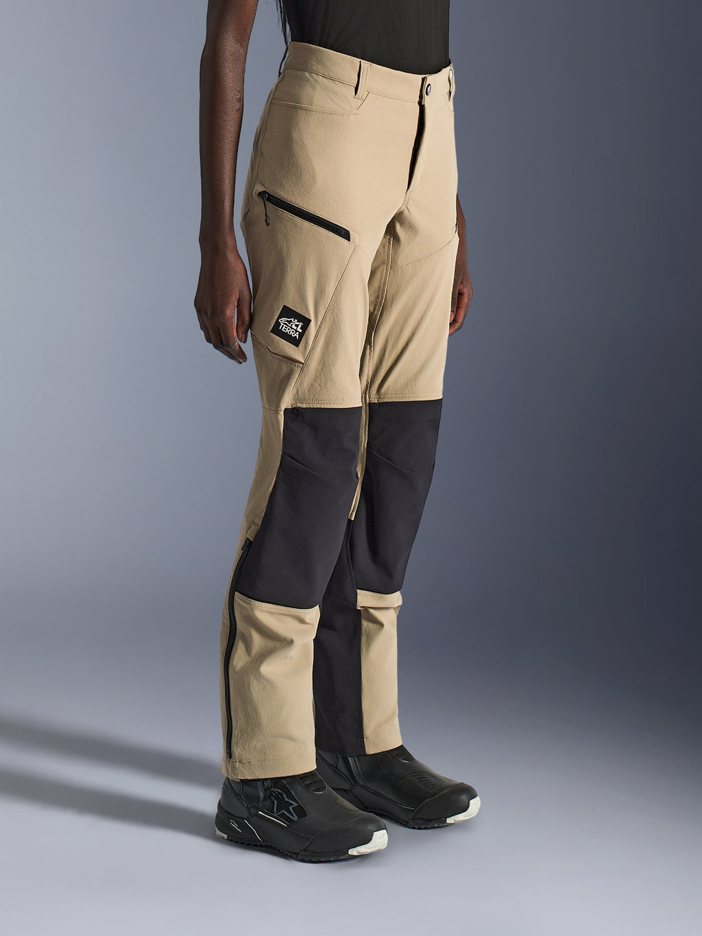 Woman wearing Alpinestars Stella Flex-AST Explorer Pants, Sand and Black adventure motorcycle pants with black ripstop knee panels, zippered cargo pockets, 'ALL TERRA' logo patch, and lower leg zippers.