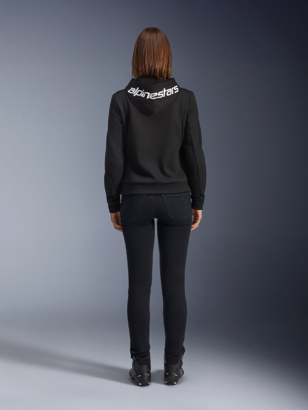 Alpinestars Daisy V3 kørejeans til damer, Black Rinse, slim fit motorcykelbukser, set bagfra med fokus på den høje talje og feminine silhuet, kombineret med en sort Alpinestars hættetrøje og sorte kørestøvler, urbant og afslappet køreudstyr
