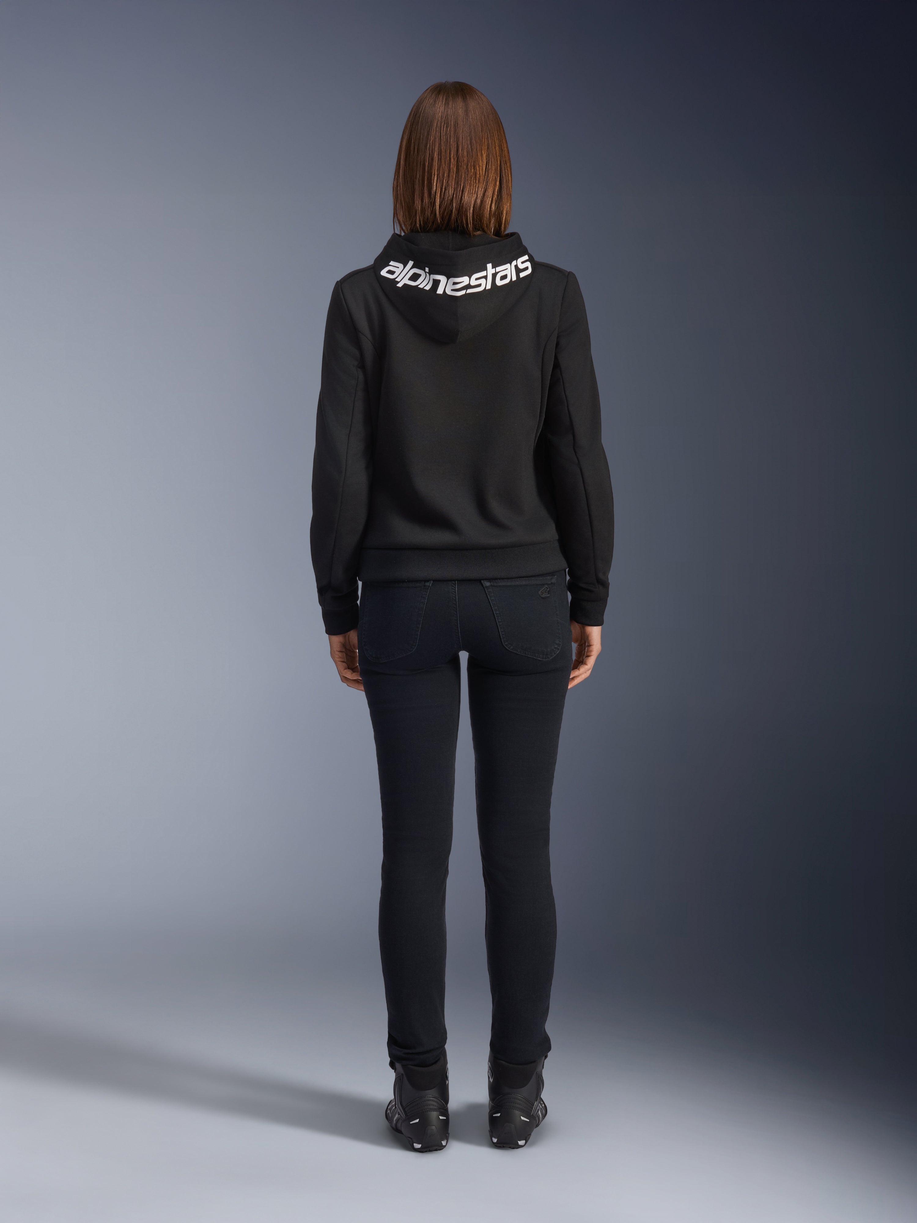 Alpinestars Daisy V3 kørejeans til damer, Black Rinse, slim fit motorcykelbukser, set bagfra med fokus på den høje talje og feminine silhuet, kombineret med en sort Alpinestars hættetrøje og sorte kørestøvler, urbant og afslappet køreudstyr