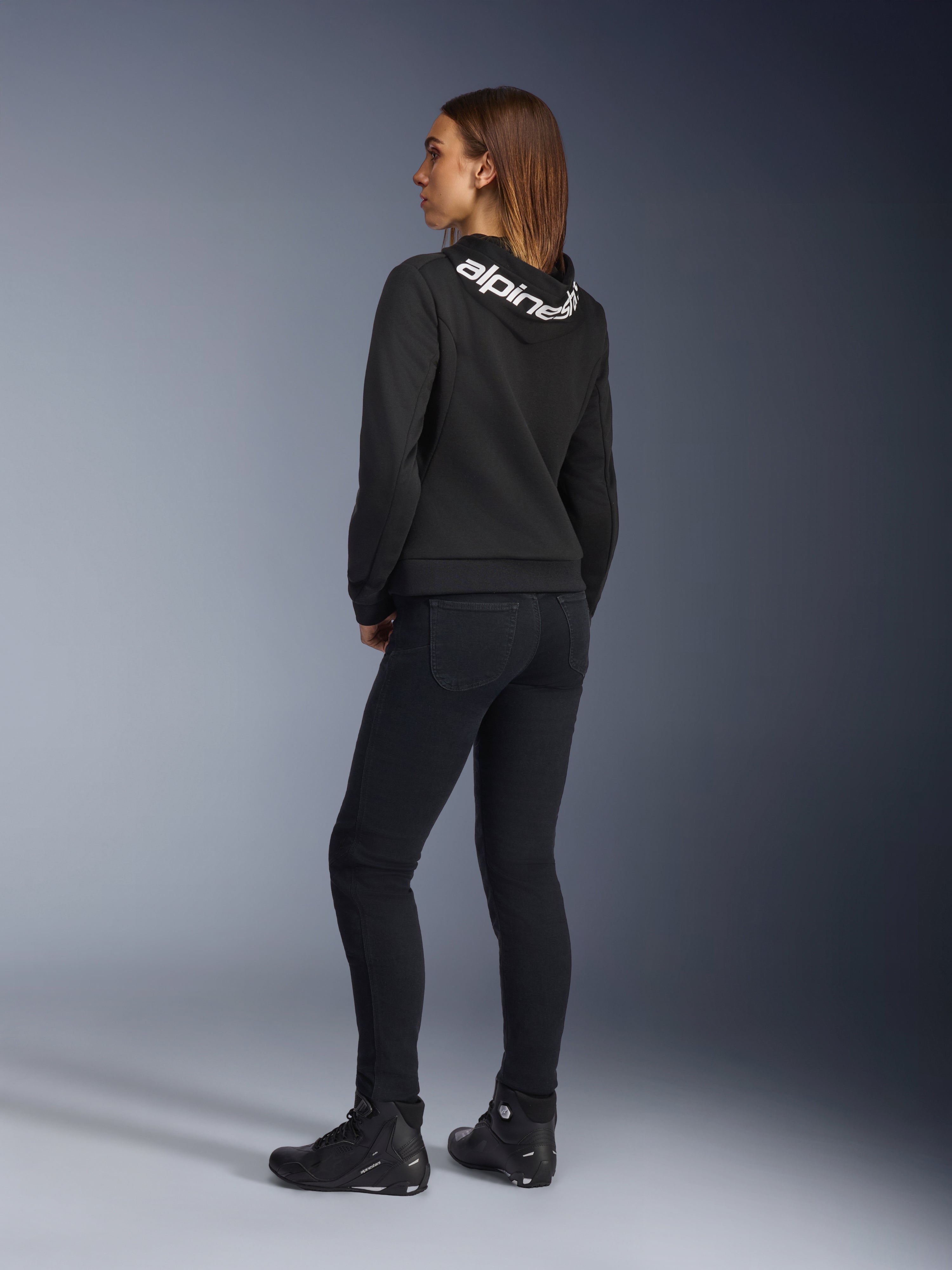 Alpinestars Daisy V3 kører-jeans til damer, motorcykelbukser til kvinder, Rinse Black, set bagfra med slim fit og høj talje bagpå, kombineret med en sort Alpinestars hættetrøje og kørestøvler for et urbant motorcykel-look