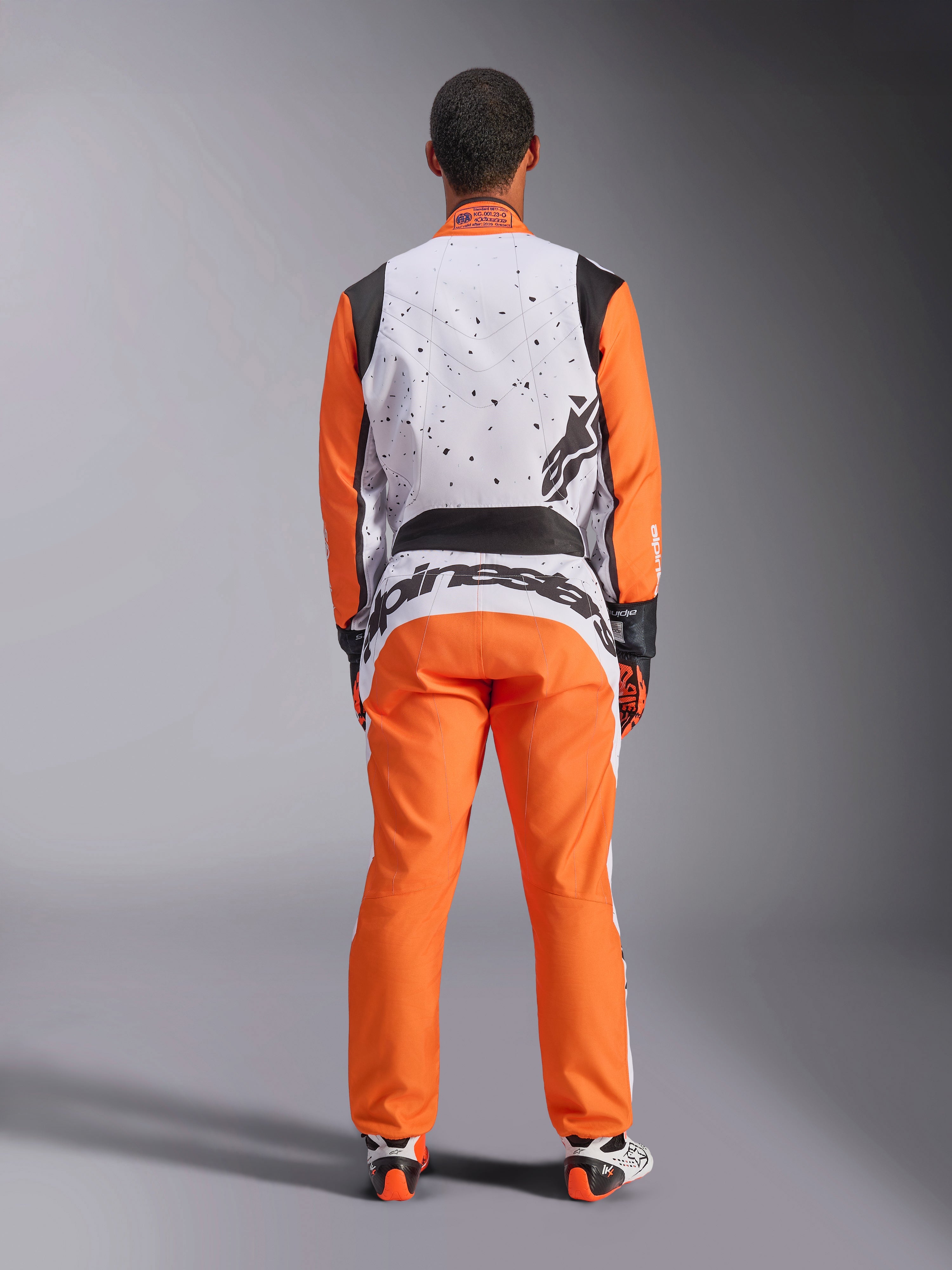 Set bagfra af en racerkører iført Alpinestars KMX-9 V3 Graphic 6 dragt i hvid, orange og sort, hvid øvre ryg med sort stænk-grafik, orange ærmer og bukser, stort sort Alpinestars-logo på lænden, kombineret med kørehandsker og støvler til gokart.