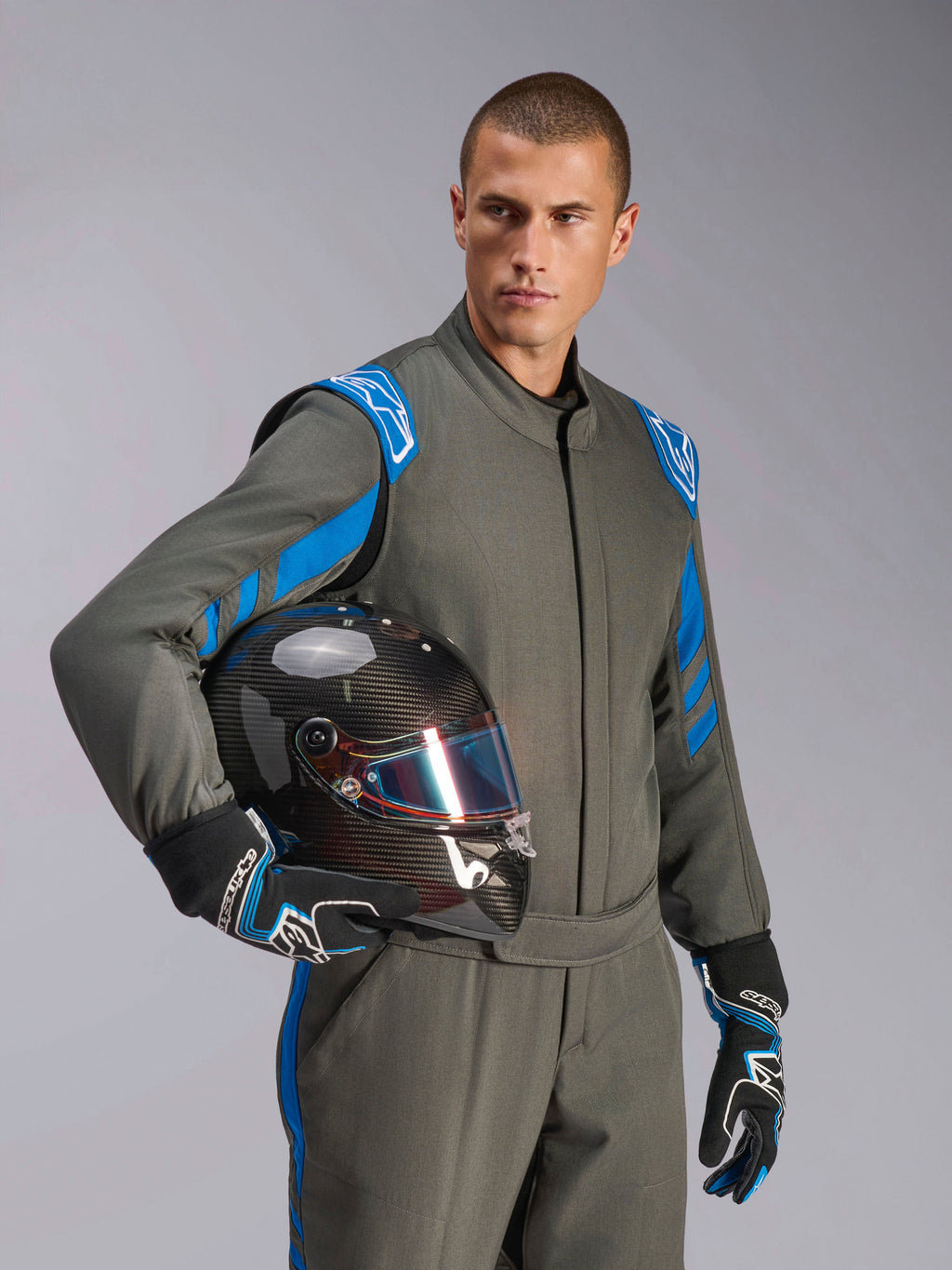 Mandlig racerkører iført Alpinestars GP Race V3 Suit FIA i Anthracite Blue, holder en kulfiber-racerhjelm med et iriserende visir, professionelt bilsportsudstyr med blå detaljer på skuldrene og matchende kørehandsker.