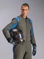 Mandlig racerkører iført Alpinestars GP Race V3 Suit FIA i Anthracite Blue, holder en kulfiber-racerhjelm med et iriserende visir, professionelt bilsportsudstyr med blå detaljer på skuldrene og matchende kørehandsker.