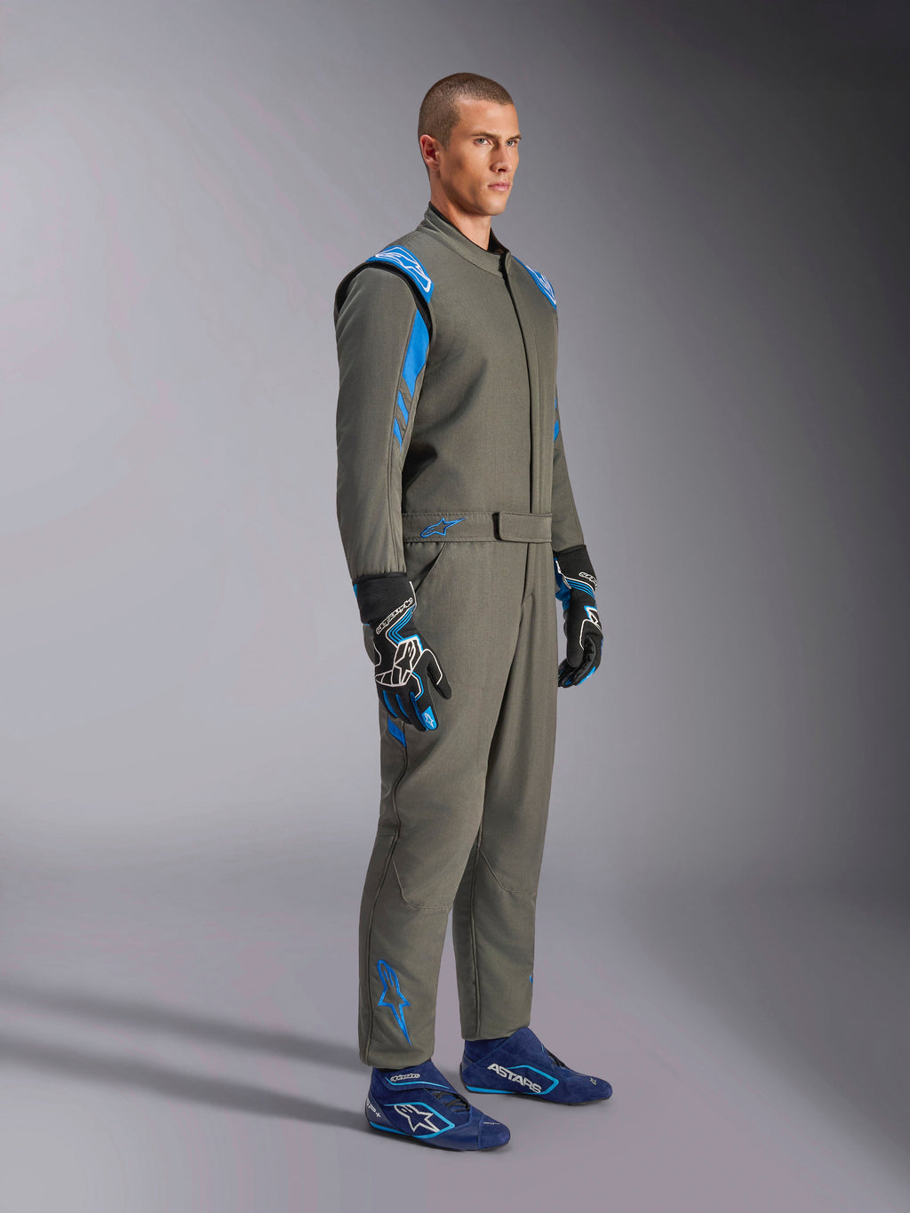 Alpinestars GP Race V3-køredragt FIA i Anthracite Blue, ét-delt autoracerdragt båret af en mandlig model, med klare blå detaljer på skuldre og ankler, kombineret med matchende racerhandsker og blå racersko, professionelt motorsportsudstyr.