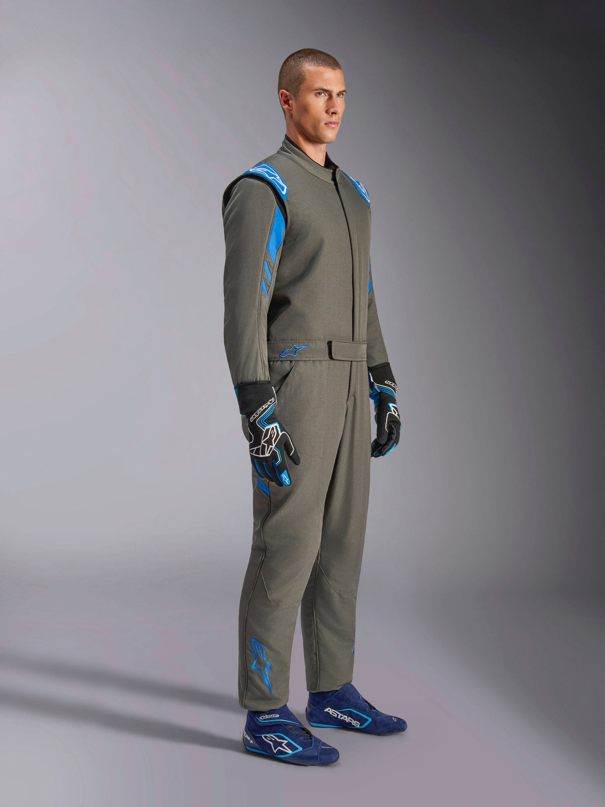 Alpinestars GP Race V3-køredragt FIA i Anthracite Blue, ét-delt autoracerdragt båret af en mandlig model, med klare blå detaljer på skuldre og ankler, kombineret med matchende racerhandsker og blå racersko, professionelt motorsportsudstyr.