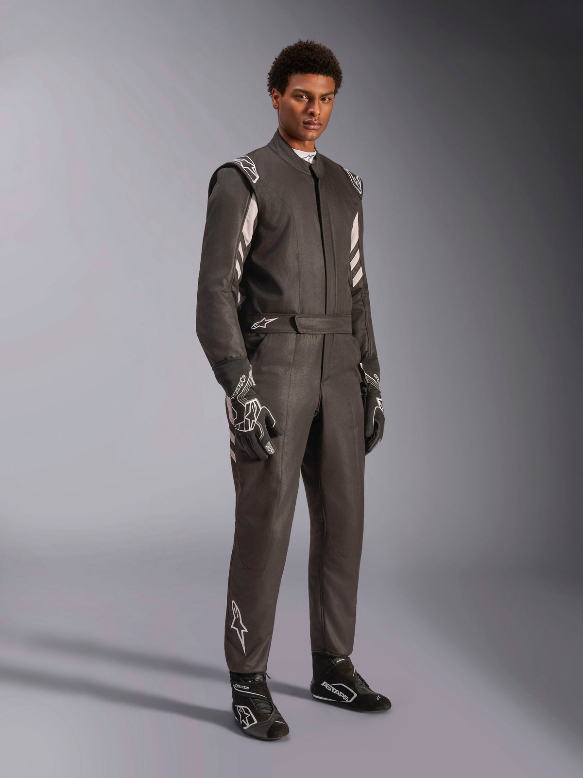 Mandlig racerkører iført Alpinestars GP Race V3 Suit FIA, sort og sølvfarvet køredragt til bilsport, komplet med matchende handsker og sko, sølvdetaljer på skuldre og underben, professionelt racerudstyr med skræddersyet pasform