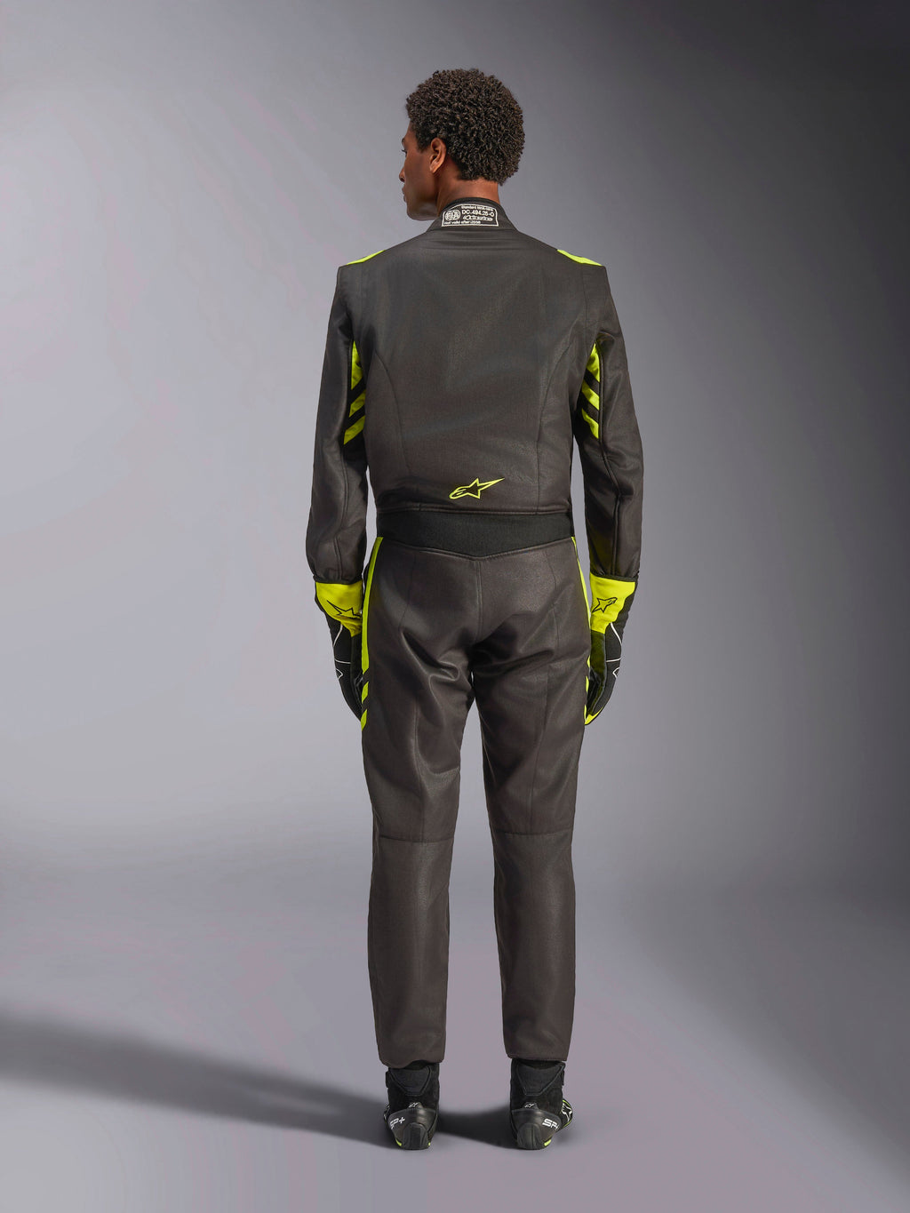 Alpinestars GP Race V3 Køredragt FIA, set bagfra, sort med neon-gule detaljer, med gule paneler på arme og overkrop, gult logo på lænden, båret med matchende racerhandsker og støvler, professionel racerdragt til bilsport.