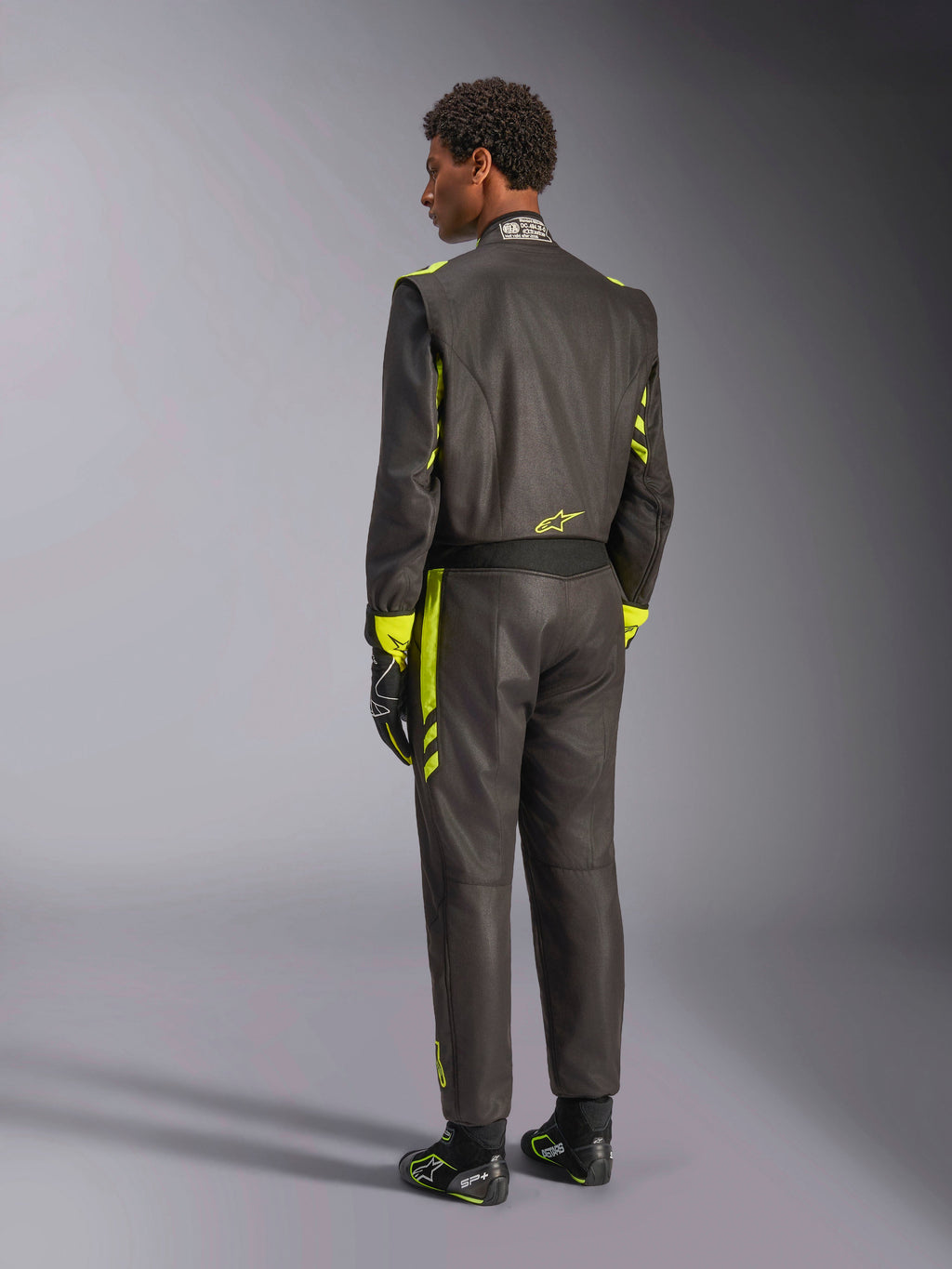 Alpinestars GP Race V3 Køredragt FIA, set bagfra, sort med neon-gule detaljer, båret med matchende kørehandsker og støvler, professionelt bilsportudstyr med en tolags aramidkonstruktion