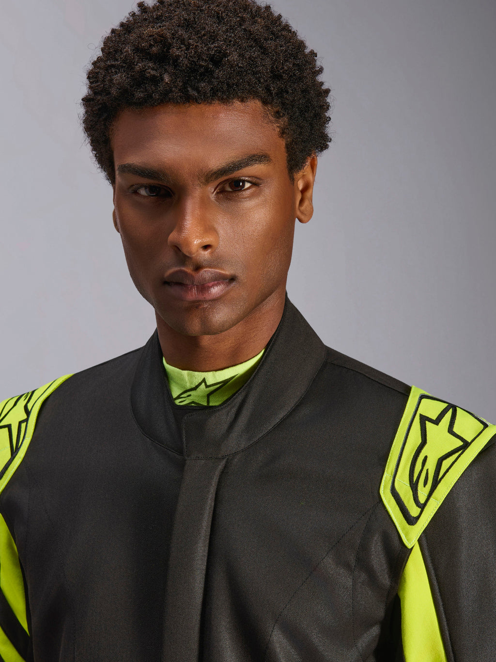 Nærbillede af en model iført Alpinestars GP Race V3 Suit FIA i sort og gul fluo, med gule udtræksepauletter på skuldrene med stjernelogoer, design med høj krave, professionelt bilsportsudstyr.