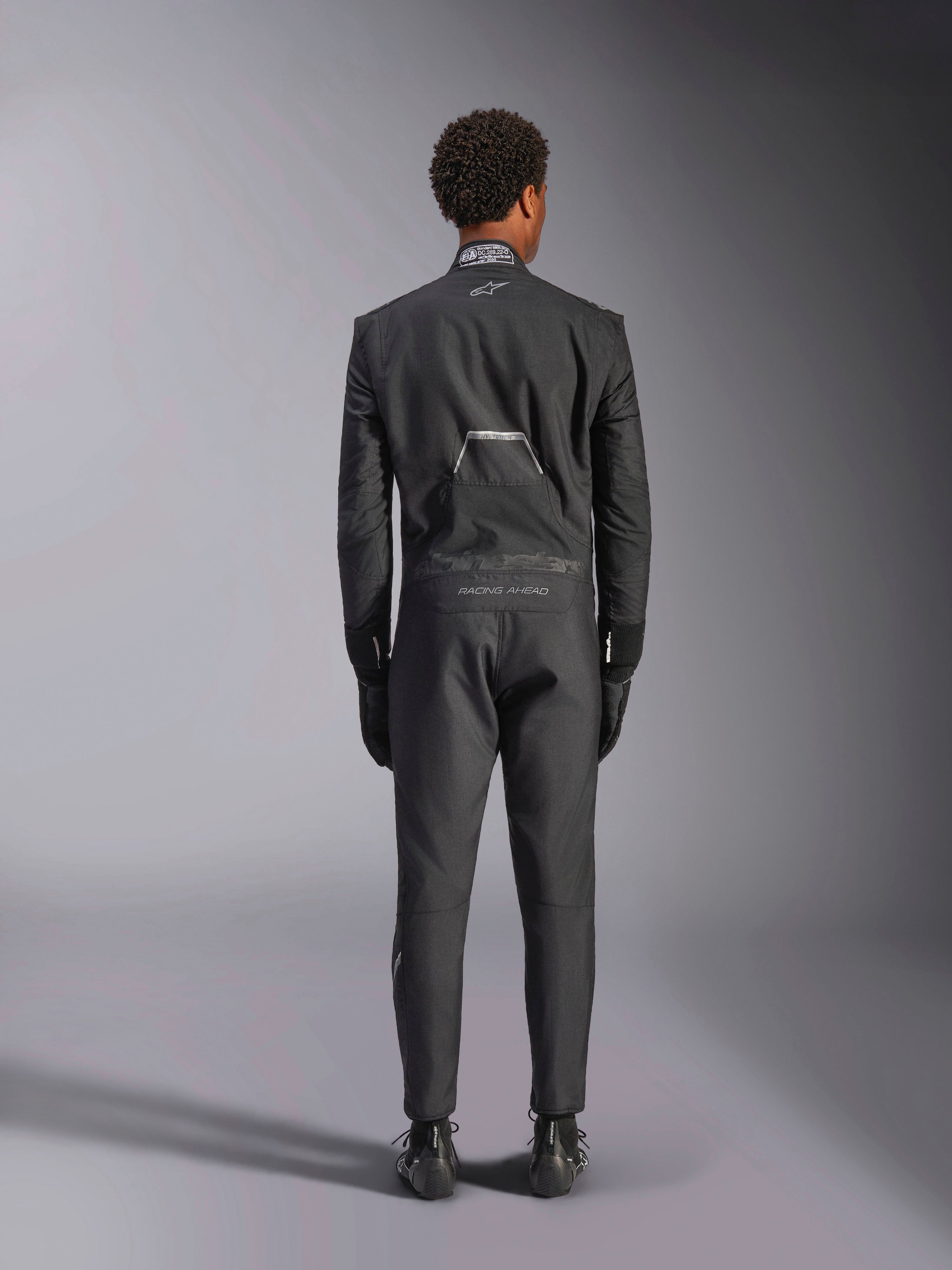 Alpinestars Hypertech V3 Suit, Black Dark Shadow, rear view of a professional one-piece auto racing suit, featuring FIA certification label on the collar and 'Alpinestars' and 'RACING AHEAD' branding on the lower back, worn with matching black racing gloves and boots.