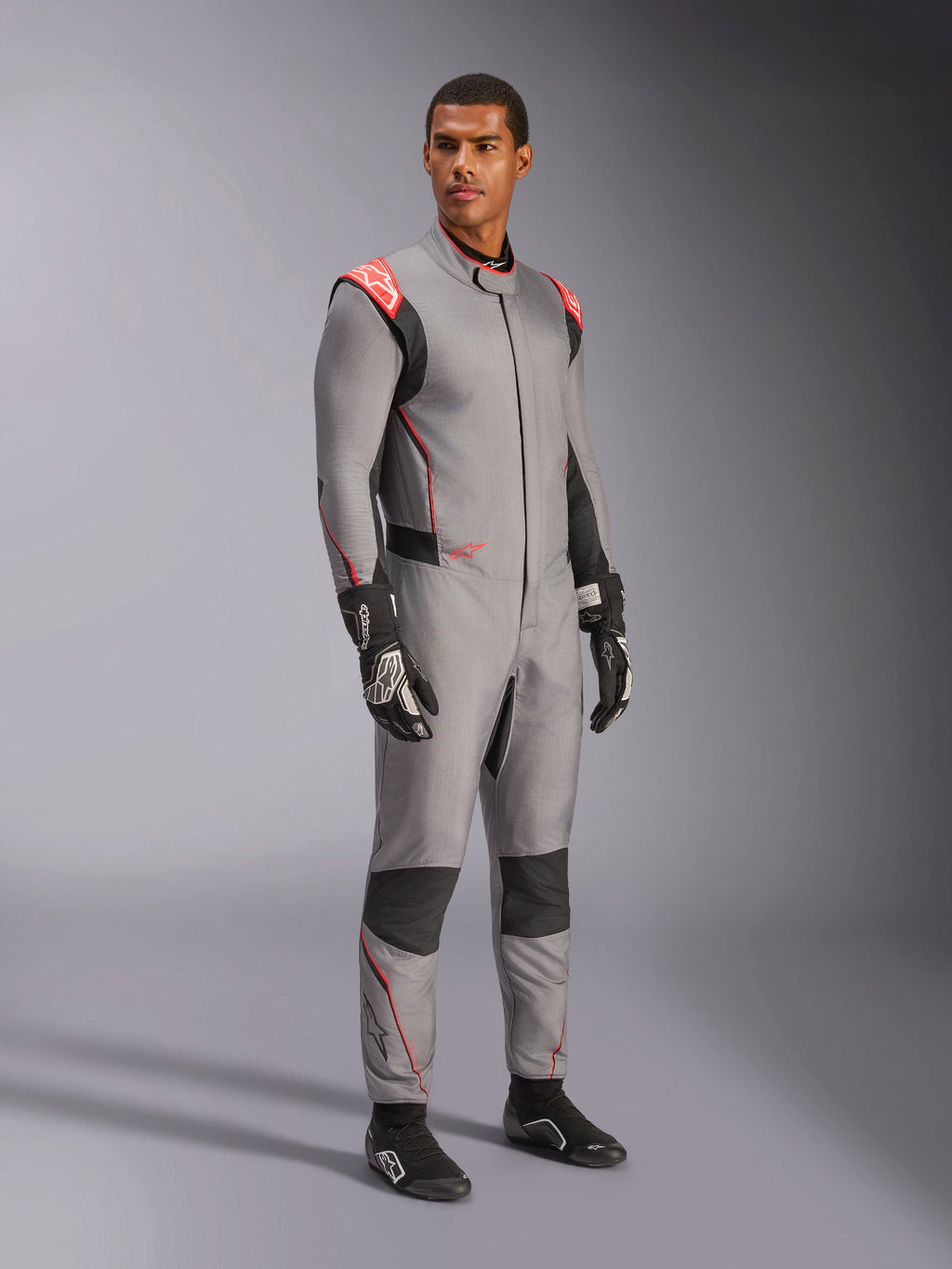 Mandlig model iført Alpinestars Hypertech V3-dragt i Mid Gray med sorte og rød fluo-detaljer, professionel autoracerdragt kombineret med sorte racerhandsker og -sko, med en letvægts to-lags Nomex-konstruktion, røde skulderdetaljer og en atletisk pasform