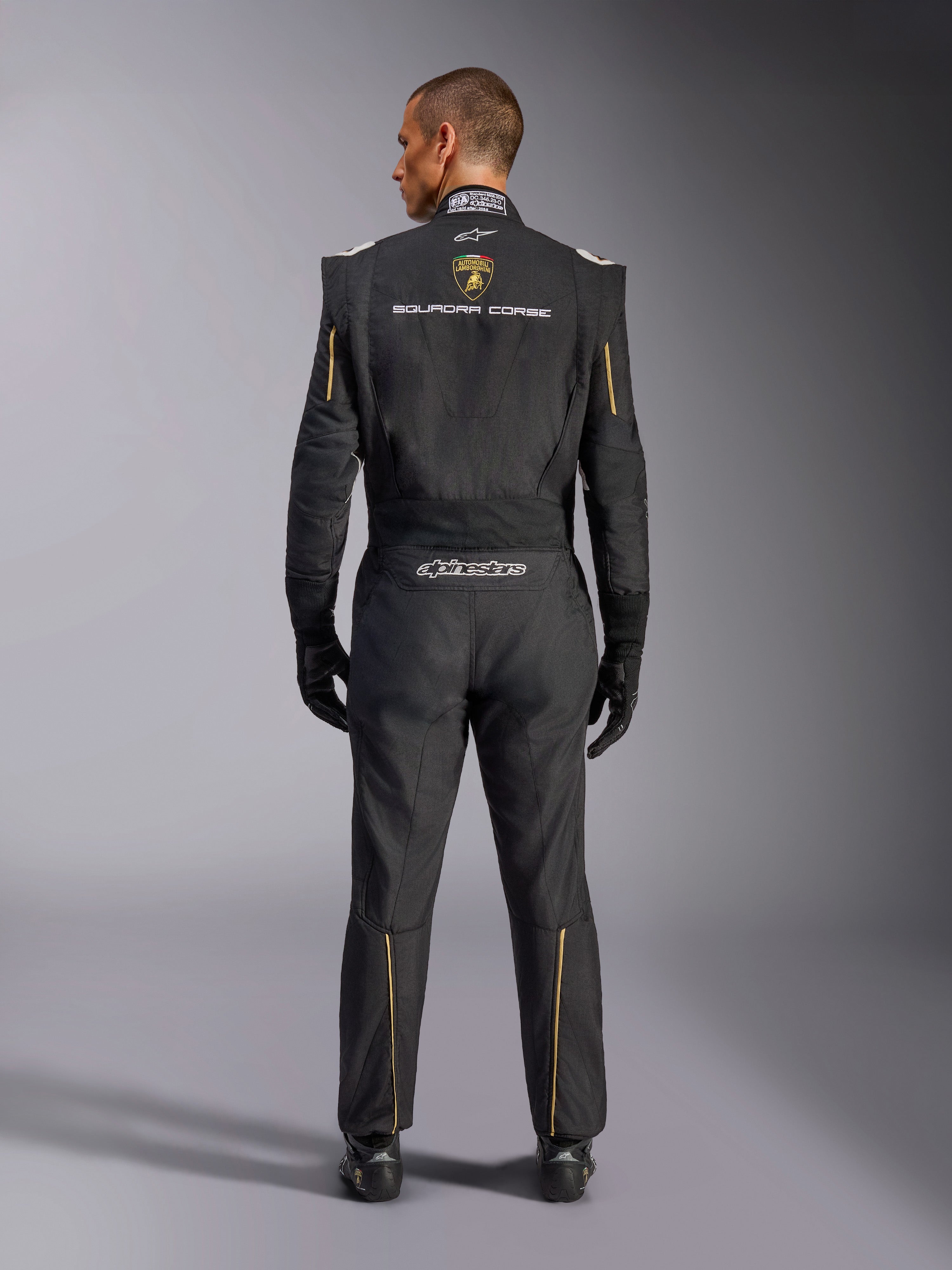 Alpinestars Limited Edition Lamborghini GP Tech V4 Suit FIA, Auto Racing Suit, Black with Gold and Silver accents, rear view showing Lamborghini Squadra Corse branding, Alpinestars logo, and gold piping on sleeves and legs, professional racing gear