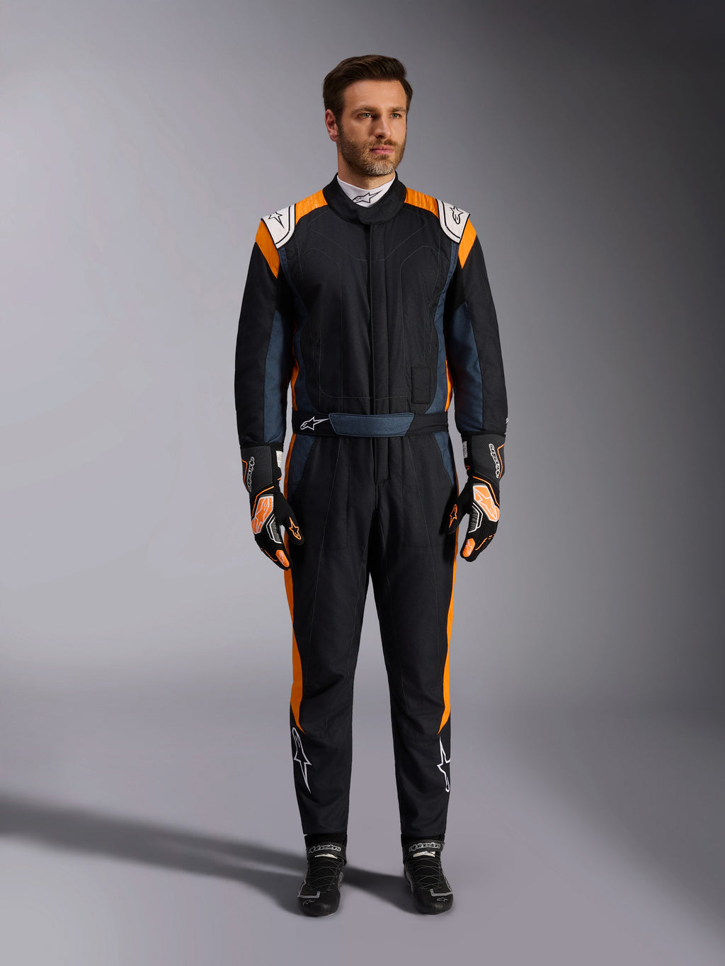 Alpinestars GP Pro Comp V2 FIA-dragt, Black Asphalt og Orange Fluo autoracerdragt, mandlig model iført fuldt beskyttelsesudstyr med matchende racerhandsker og -sko, hvide Alpinestars-logoer og markante orange sidepaneler, professionelt motorsportstøj
