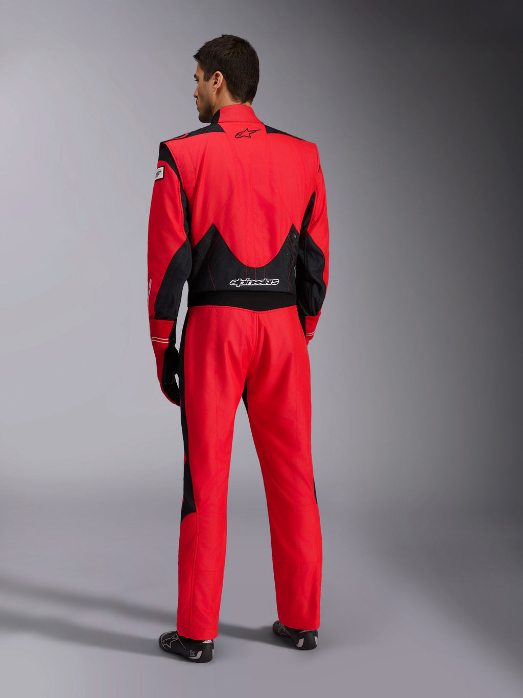 Alpinestars GP Pro Comp V2 FIA køredragt set bagfra, rød og sort, med åndbare sorte mesh-paneler på skuldrene og lænden, hvidt Alpinestars-logo, bæres med matchende racerhandsker og støvler, professionelt motorsportsudstyr