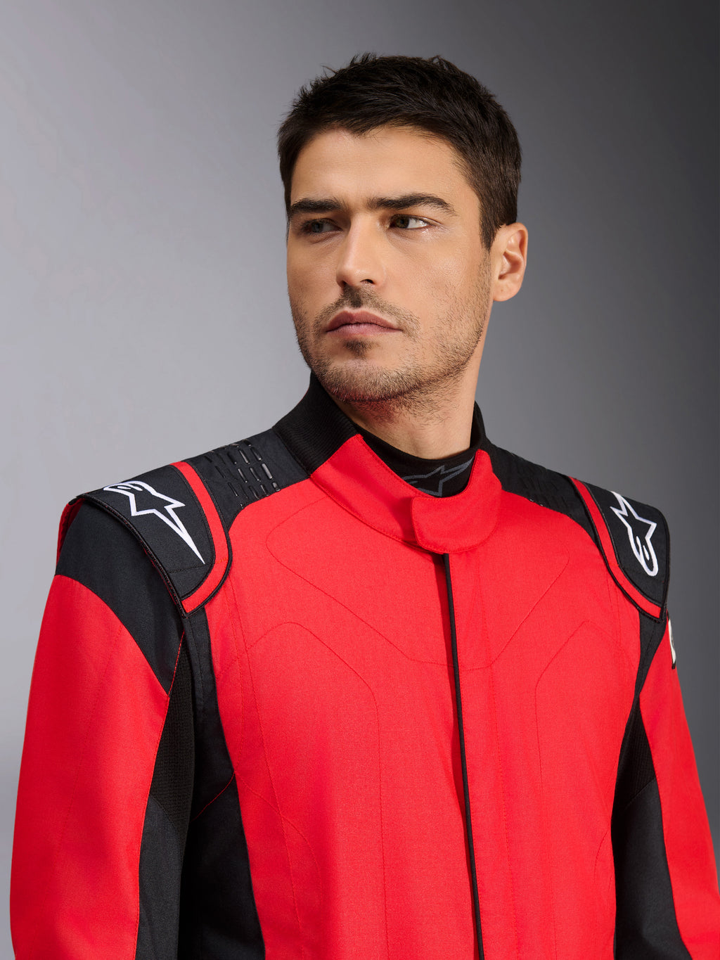Mandlig model iført Alpinestars GP Pro Comp V2 FIA køredragt, rød og sort, med hvide Astars-logoer på skuldrene og detaljer med silikonegrebsteknologi, professionel beklædning til motorsport