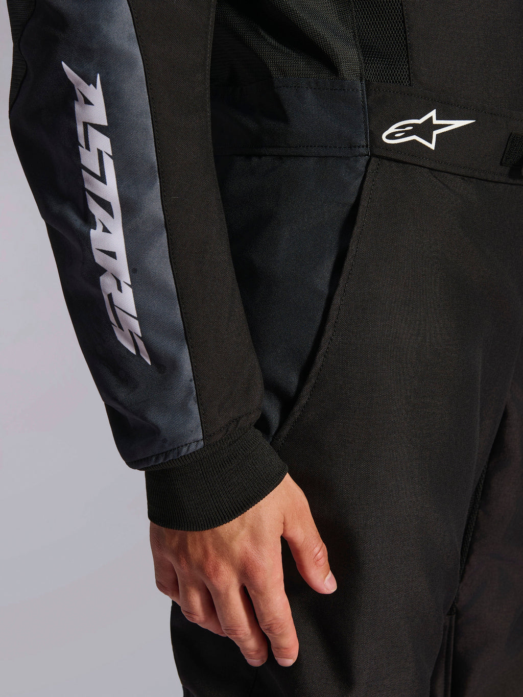 Alpinestars KMX-5 V3-dragt, kartingdragt, sort og Dark Shadow, nærbillede af detalje på hofte og arm, hvid ASTARS-branding på ærmet, hvidt logo i taljen, med mesh-paneler til ventilation og en strikket manchet ved håndleddet