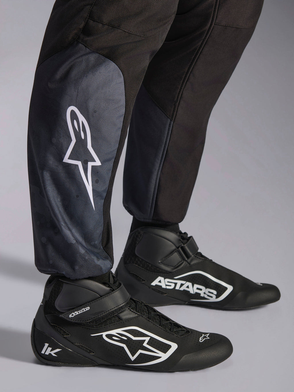 Alpinestars KMX-5 V3 dragt, Black Dark Shadow, nærbillede af underben og ankler, hvidt stjernelogo på læggen, parret med sorte kartingsko, fremhæver strikkede ankelmanchetter og atletisk-regulær pasform.