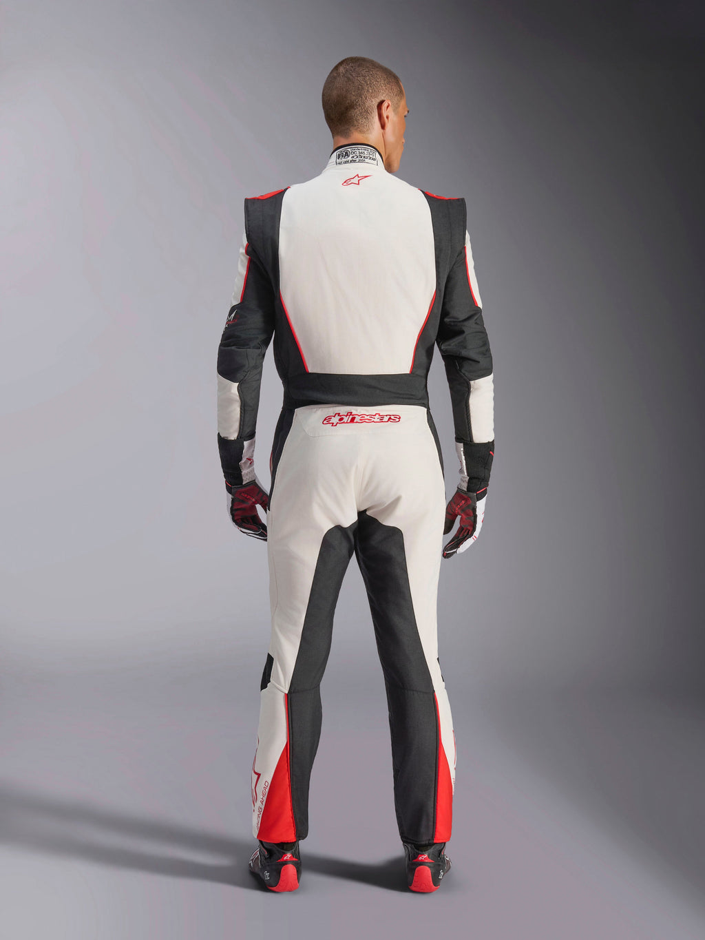 Alpinestars GP Tech V4 Suit, Auto Racing Suit, White Black and Red, back view of driver wearing the one-piece suit, featuring black stretch panels on the sides and shoulders, red accents on the lower legs, Alpinestars branding on the lower back and collar, professional FIA-certified motorsport gear