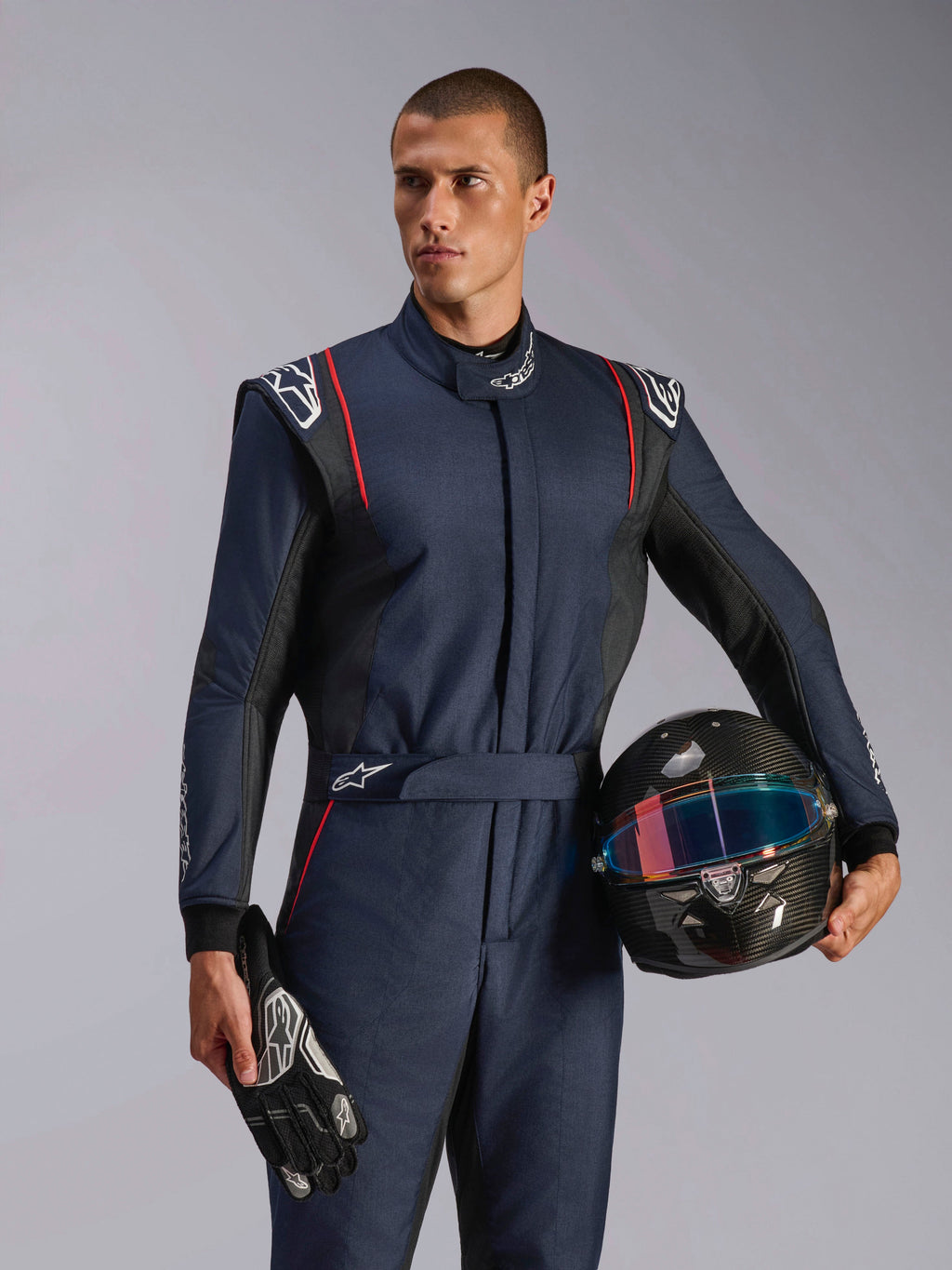 Male model wearing Alpinestars GP Tech V4 Suit, Navy Blue auto racing suit with black and red accents, holding a carbon fiber helmet and racing gloves, professional motorsport protective gear
