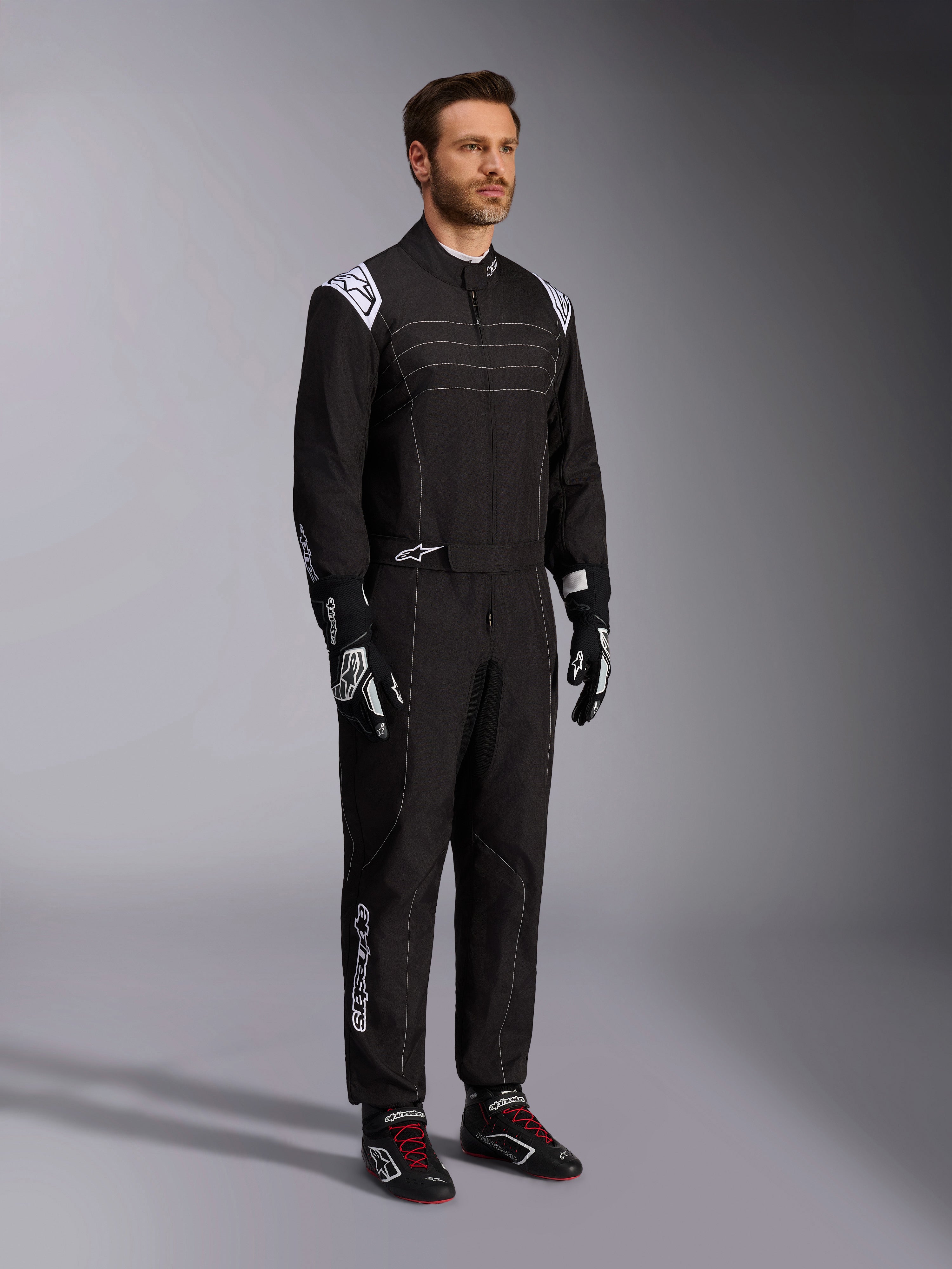 Alpinestars KMX-9 V3 kartingdragt, sort og hvid, mandlig model iført fuld racerdragt med hvidt Alpinestars-logo på skuldrene, kombineret med sort-hvide racerhandsker og sorte kartingsko med røde snørebånd, studieoptagelse der viser front- og sideprofil
