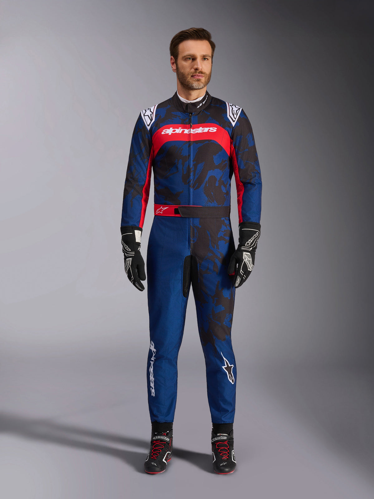 Alpinestars KMX-9 V3 Graphic 5 køredragt, kartingdragt i navyblå og rød med et abstrakt grafisk camouflage-mønster, båret af en model med sorte kørehandsker og sko, atletisk design med fremtrædende Alpinestars-branding på brystet og benene.