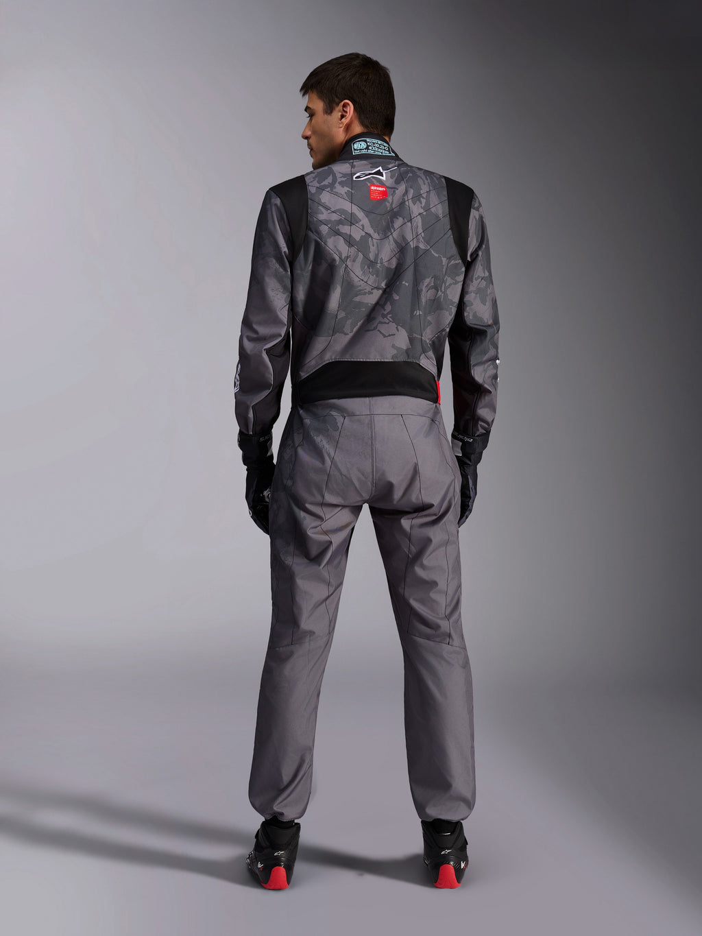 Alpinestars KMX-9 V3 Graphic 5 køredragt, kartingdragt, mørkegråt og sort kamuflagemønster på overkroppen, set bagfra med strækpaneler og Alpinestars-branding, båret med sorte handsker og sorte racersko med røde detaljer, professionelt motorsport-racerudstyr