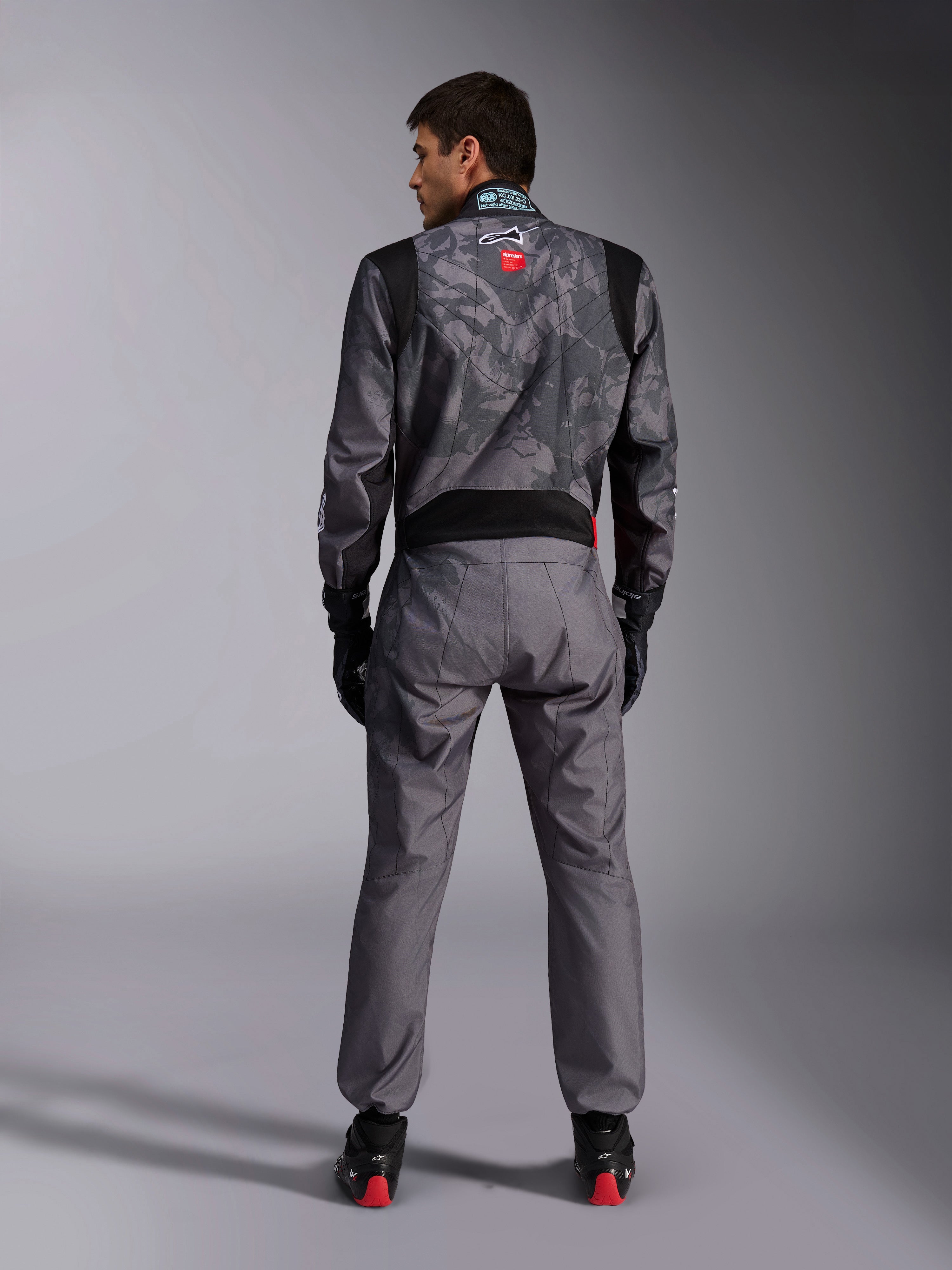 Alpinestars KMX-9 V3 Graphic 5 køredragt, kartingdragt, mørkegråt og sort kamuflagemønster på overkroppen, set bagfra med strækpaneler og Alpinestars-branding, båret med sorte handsker og sorte racersko med røde detaljer, professionelt motorsport-racerudstyr