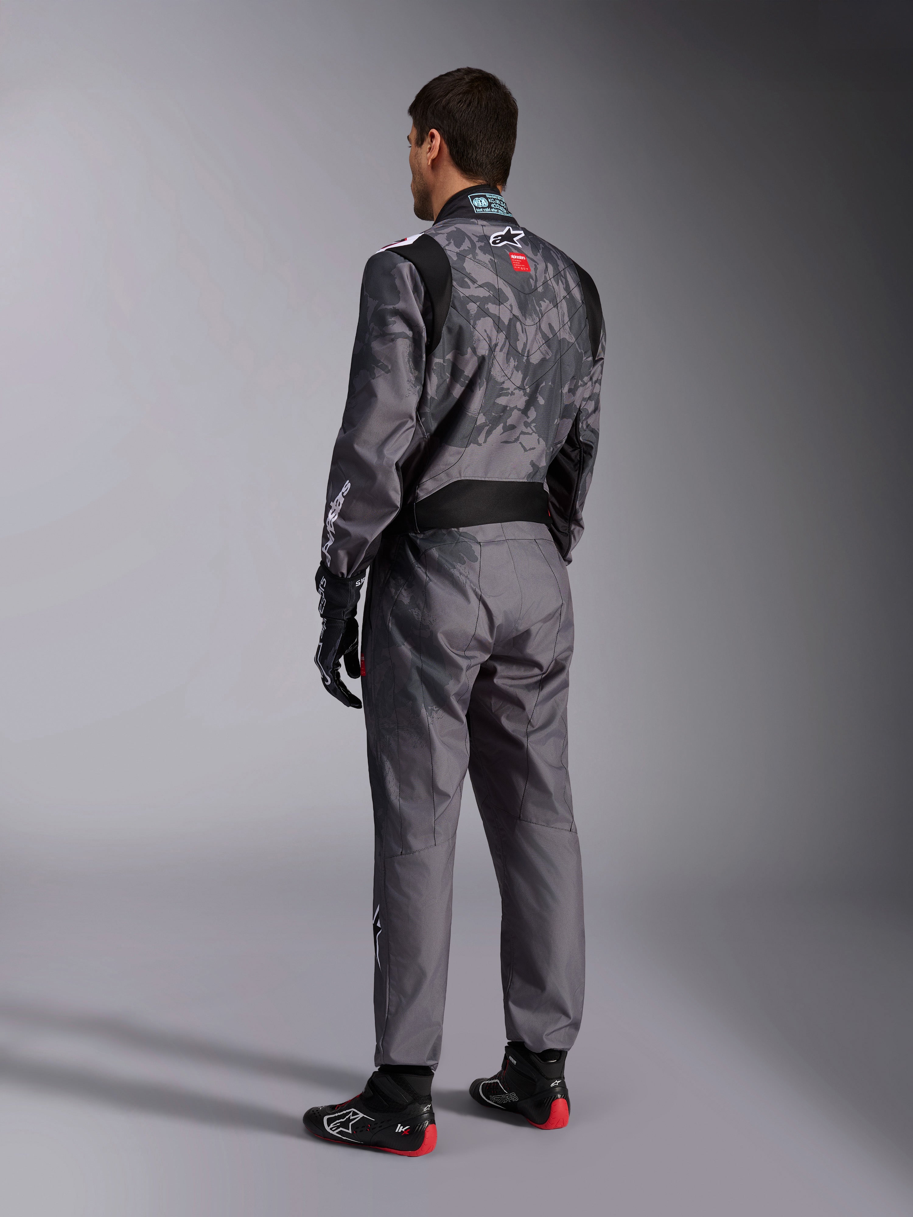 Alpinestars KMX-9 V3 Graphic 5 Suit, mørkegrå og sort kartingdragt, set bagfra af model iført komplet racersæt inklusiv sorte og røde handsker og kartingsko, med integrerede grafiske mønstre og branding på ryggen.