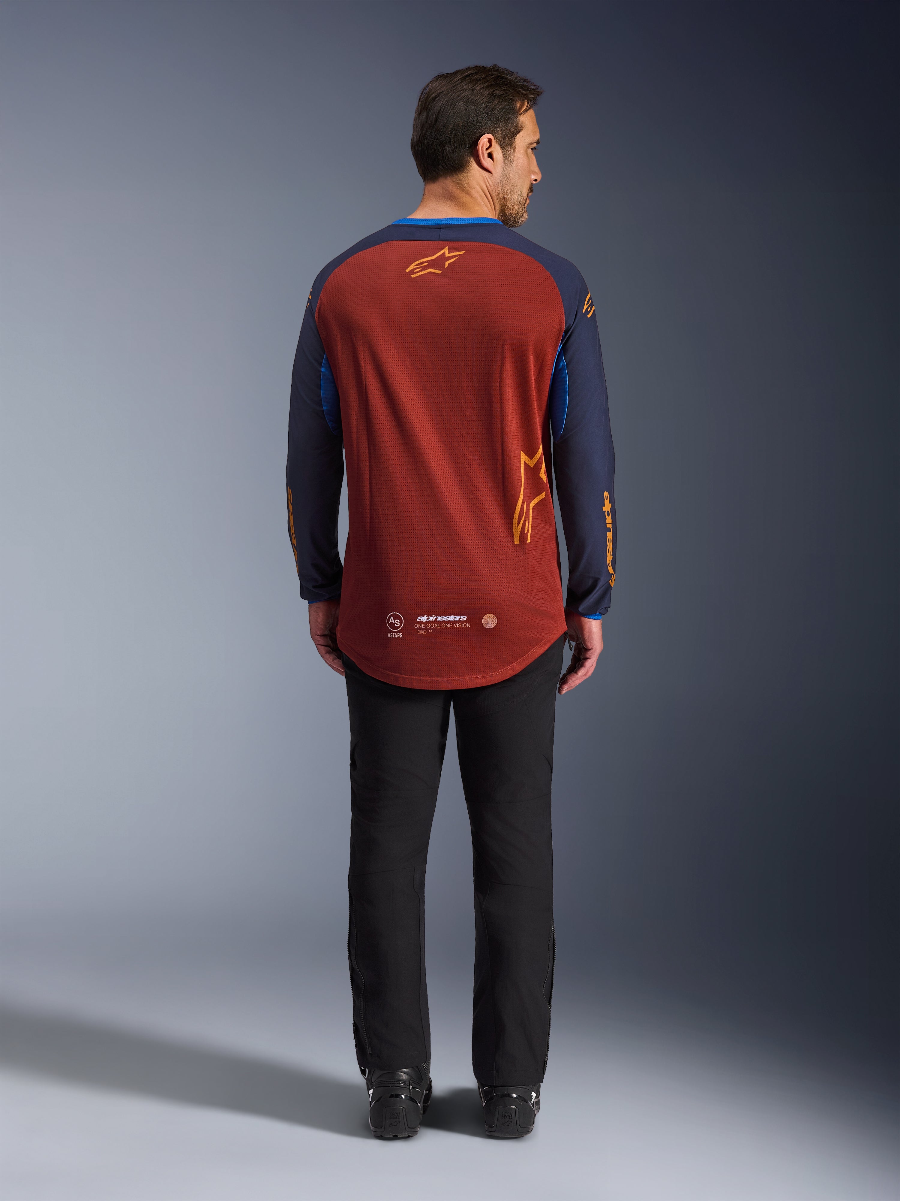 Alpinestars Maxdura Dual Jersey i Rust Brown og Mood Indigo, set bagfra, som viser den forlængede ryg og logodetaljer, båret med sorte tekniske bukser og støvler, designet til adventure- og dual-sport motorcykelkørsel.