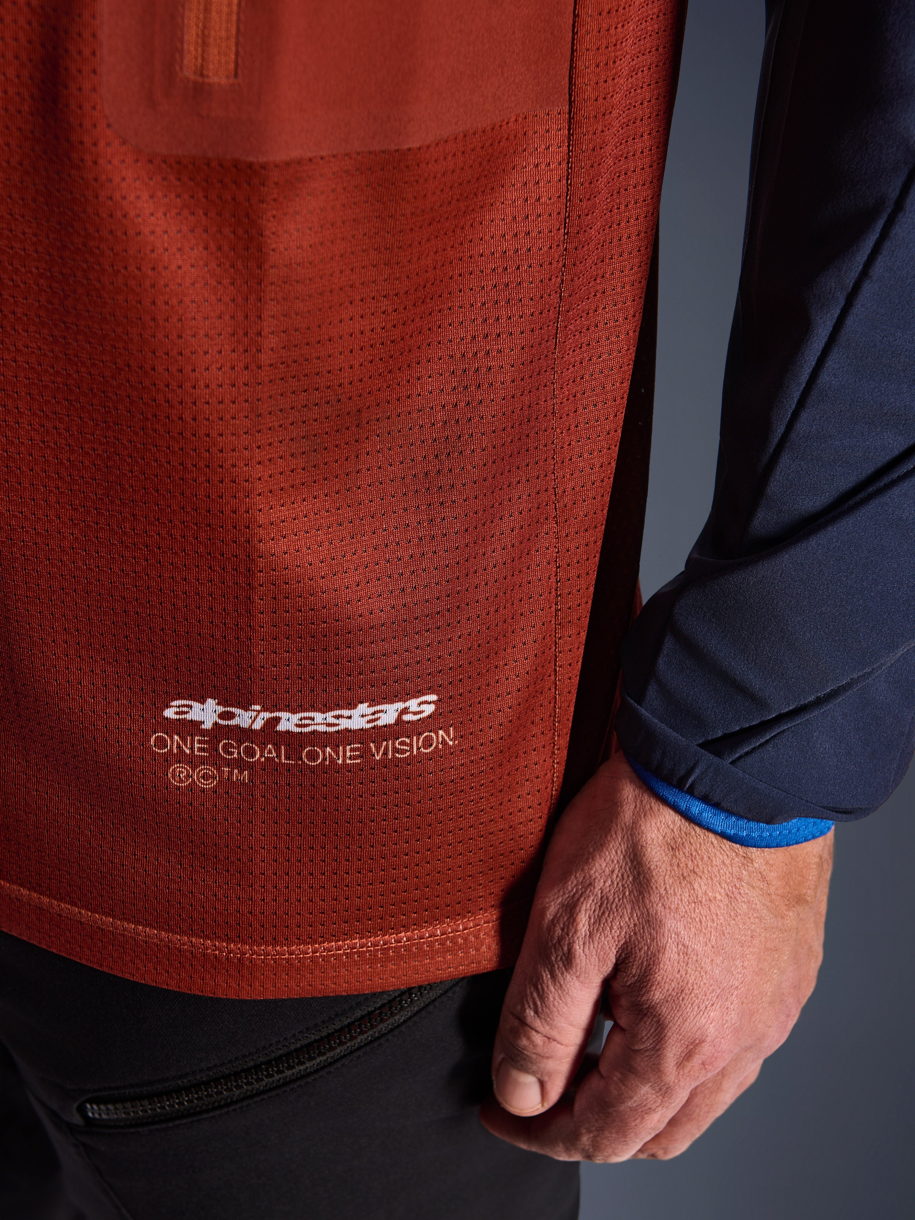 Alpinestars Maxdura Dual Jersey, adventure motorcykeltrøje, rustbrun og Mood Indigo, nærbillede af mesh-stoffet på torsoen og det mørkeblå vævede ærme, hvid Alpinestars 'One Goal One Vision' branding på den nederste søm.