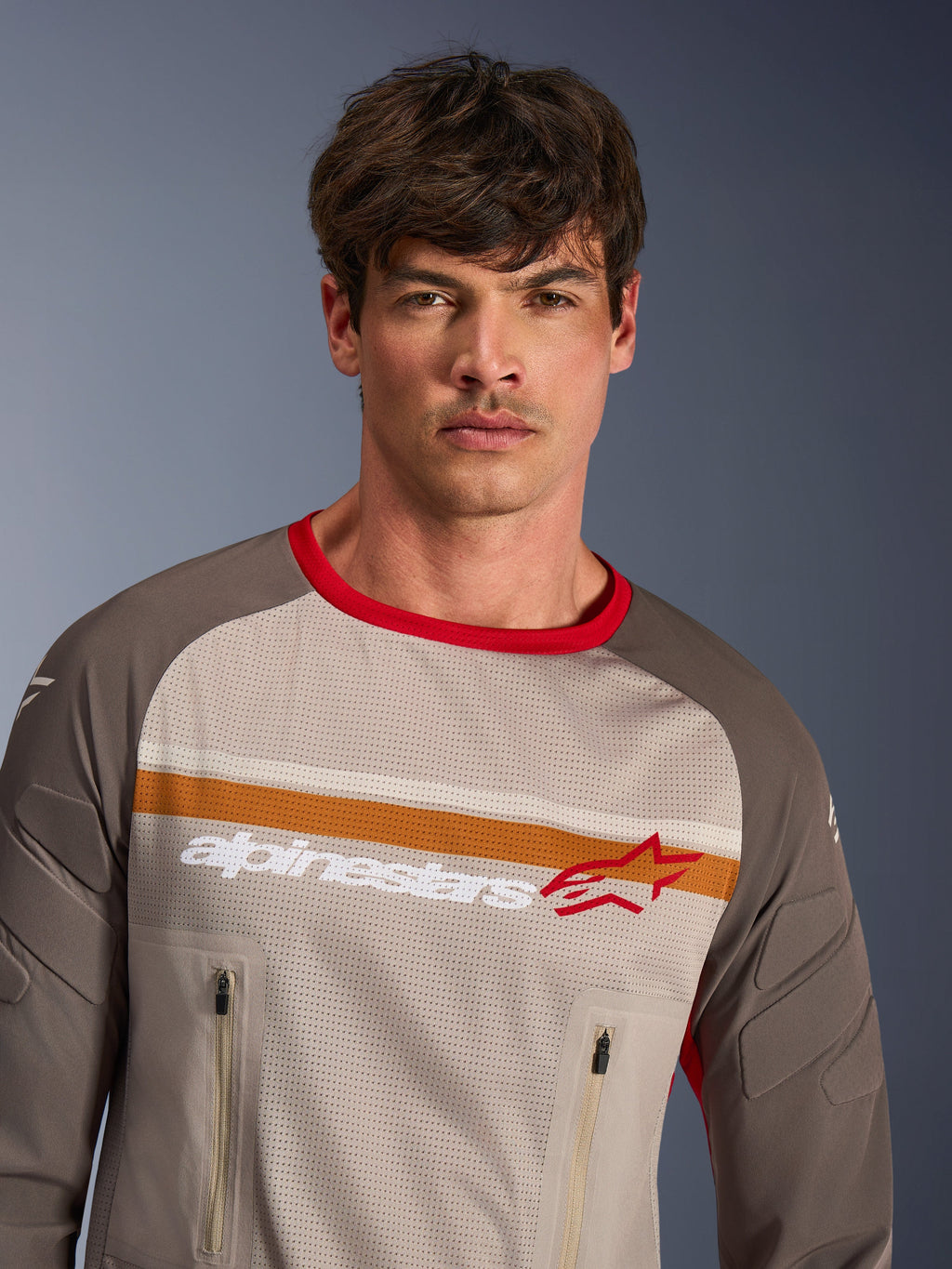 Mandlig model iført Alpinestars Maxdura Dual Jersey i Aluminum Walnut, adventure motorcykeltrøje, fuld mesh-overkrop, slidstærke ærmer, rød krave, lynlåslommer på brystet, orange og hvide striber på brystet, Alpinestars-logo