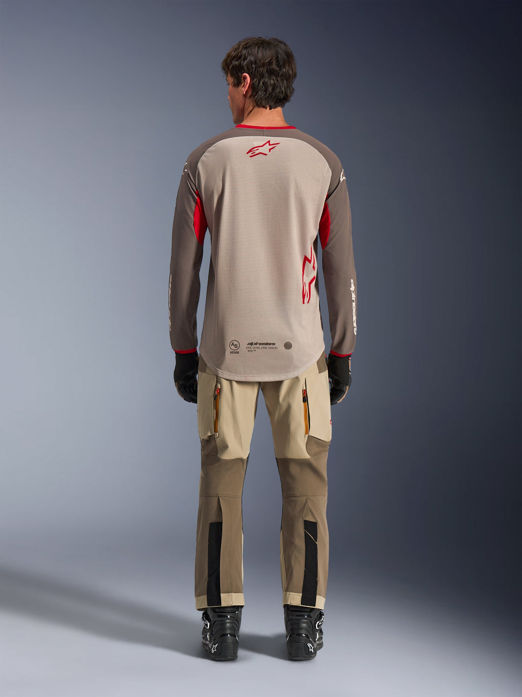 Alpinestars Maxdura Dual Jersey i Aluminum Walnut, set bagfra, beige mesh-overkrop med brune ærmer og røde logoer, forlænget bagkant, kombineret med adventure-kørebukser og sorte støvler.
