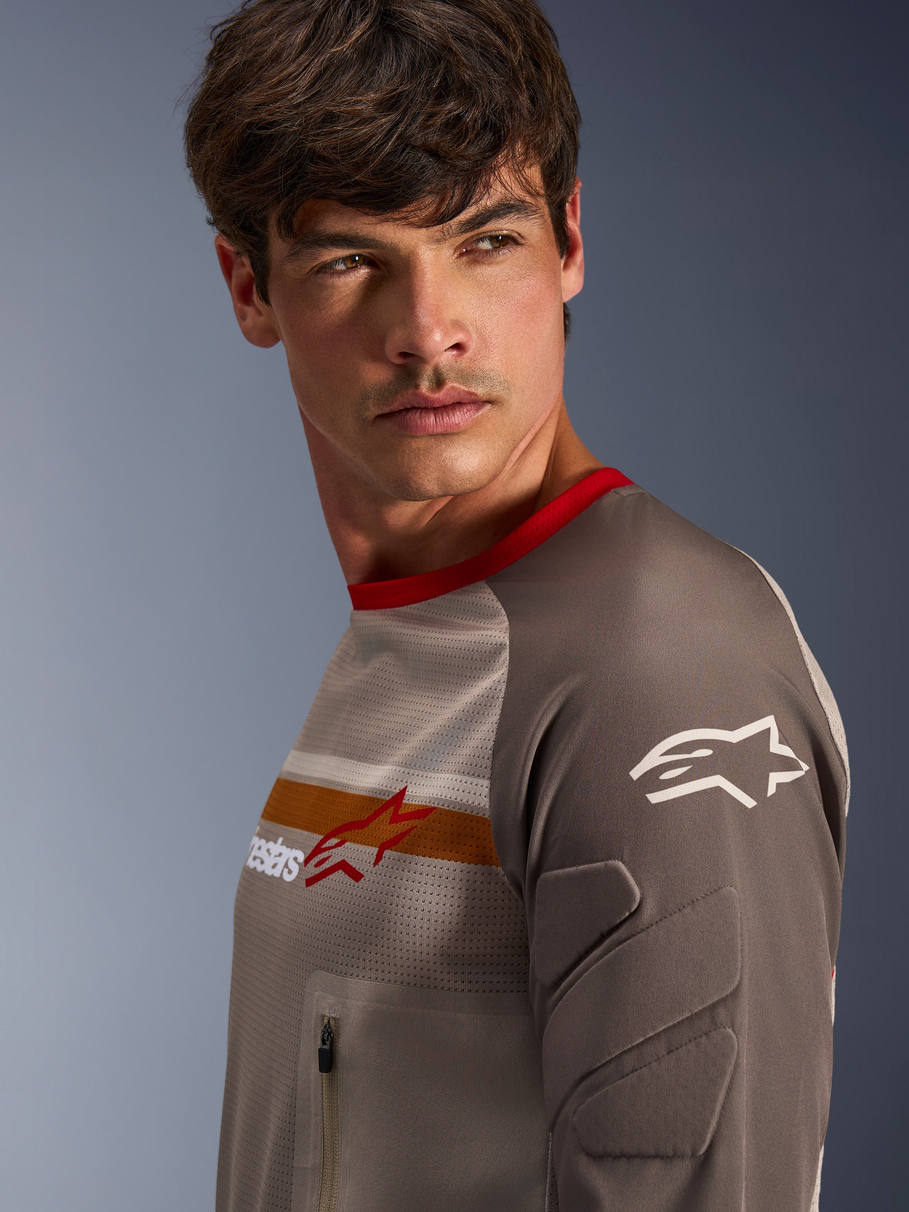 Mandlig model iført Alpinestars Maxdura Dual Jersey i Aluminum Walnut, med ventileret mesh-overkrop, rød krave og orange bryststribe. Nærbillede af slidstærkt ærme med integreret polstring og en brystlomme med lynlås til adventure-kørsel.