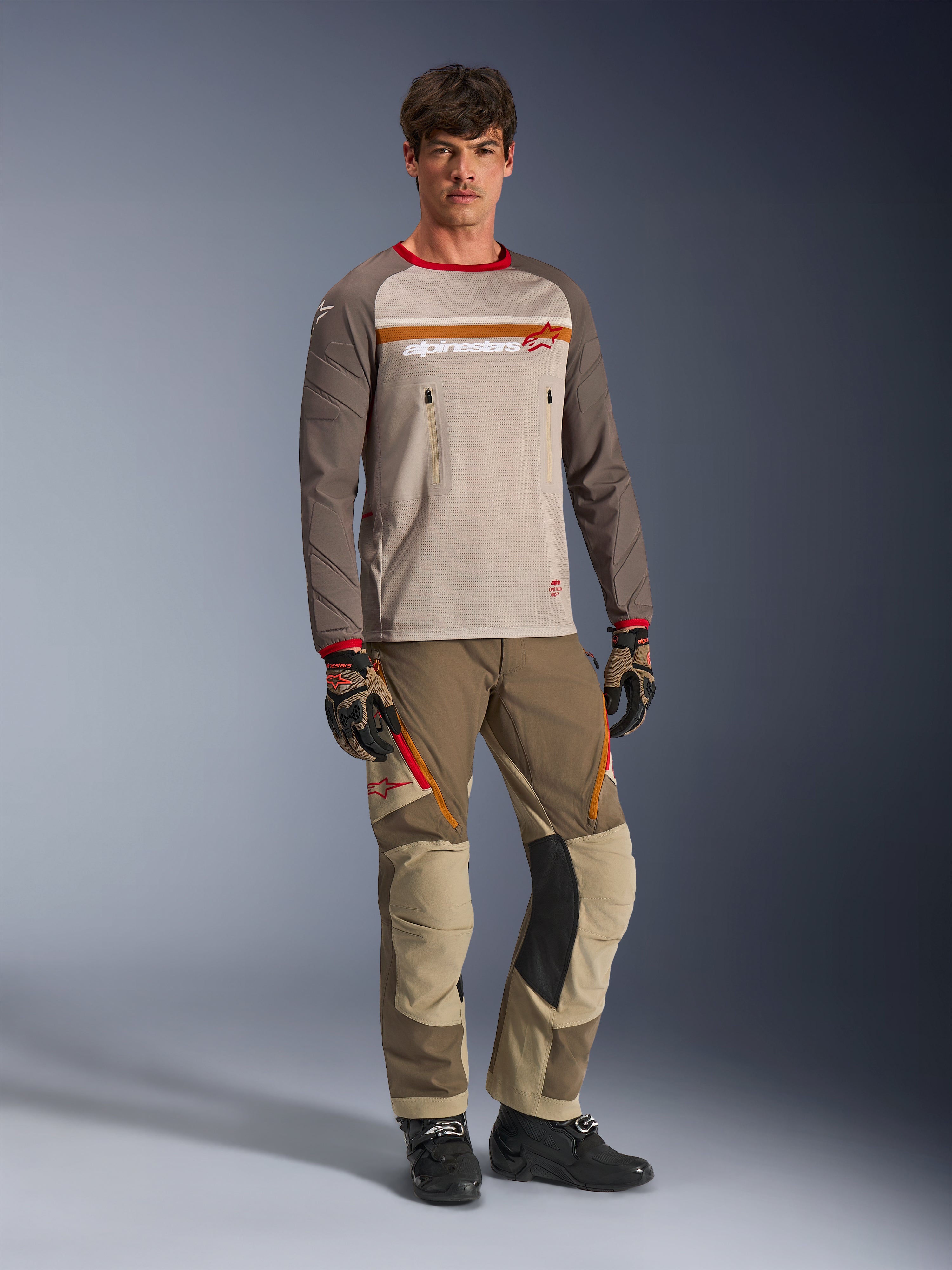 Model iført Alpinestars Maxdura Dual Jersey i Aluminum Walnut, med grå mesh-overkrop, brune ærmer med røde manchetter og brystlommer med lynlås, stylet med matchende off-road udstyr til adventure-kørsel.