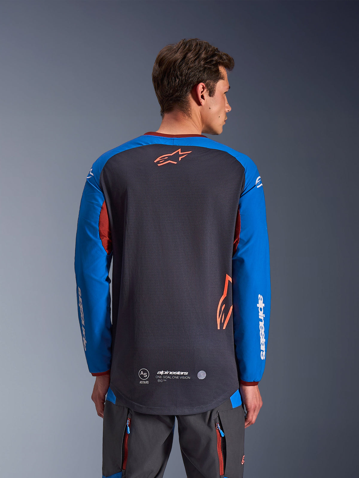 Alpinestars Maxdura Dual Jersey i Deep Gray og Royal Blue, rygvisning af mandlig model, mesh-konstruktion på torso med orange Astars-logo, lange ærmer med hvid branding, bordeauxrød krave og manchetter, båret med grå tekniske bukser, dual-sport og adventure motorcykeludstyr