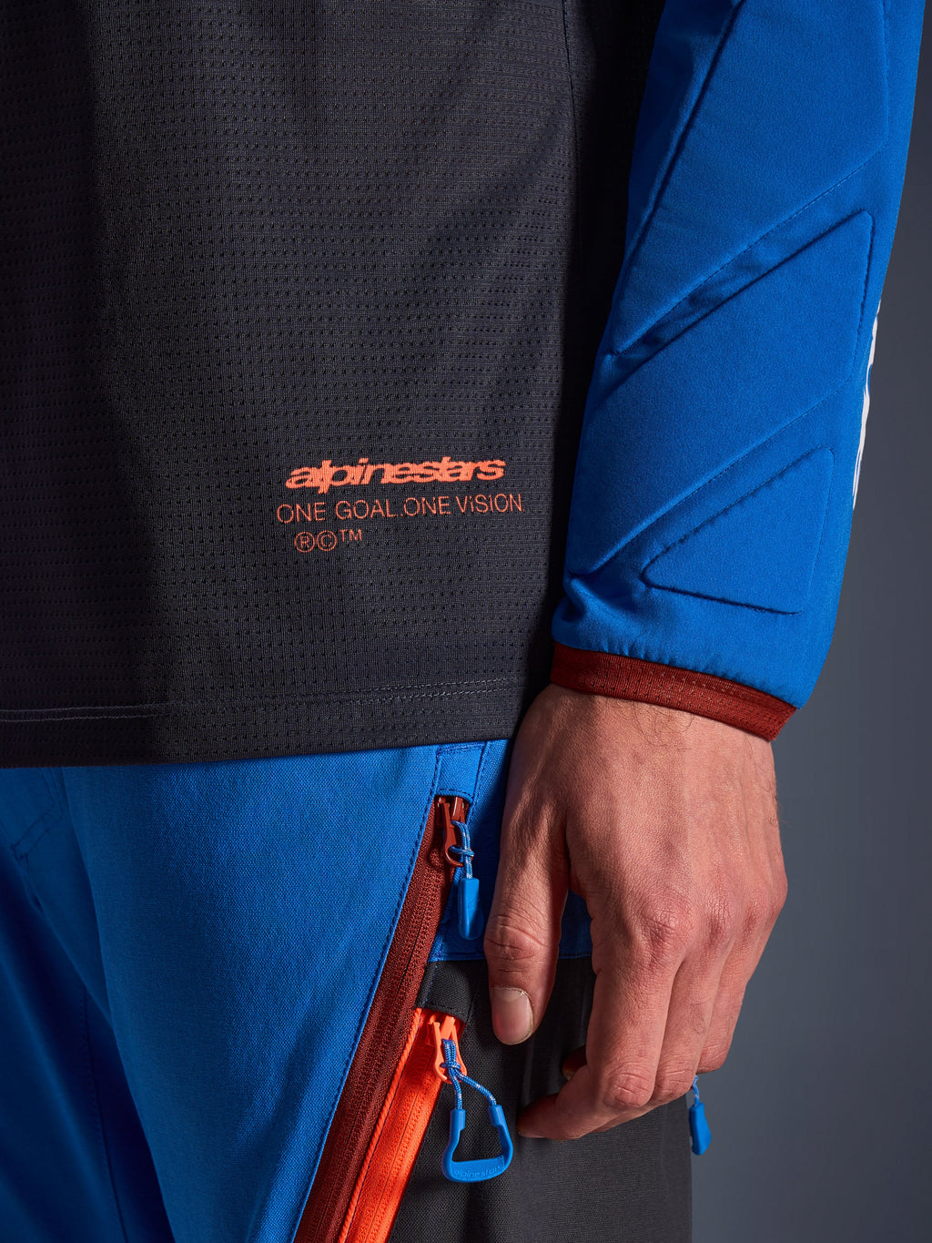 Nærbillede af Alpinestars Maxdura Dual Jersey i Deep Gray og Royal Blue, med orange logo og 'One Goal One Vision' tekst på mesh-overkroppen, polstret ærme med forstærkede geometriske paneler, teknisk adventure-køreudstyrsdetalje