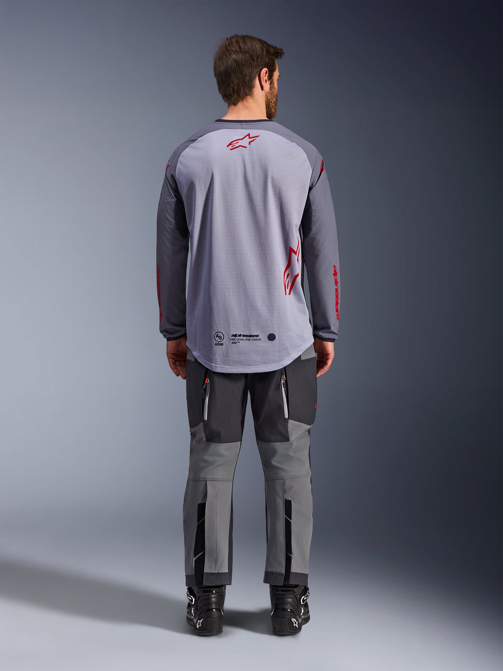Mandlig model set bagfra iført Alpinestars Maxdura Dual Jersey, mellemgrå og mørkegrå adventure-trøje med røde logo-detaljer, mesh-rygkonstruktion, båret med matchende grå off-road bukser og sorte støvler
