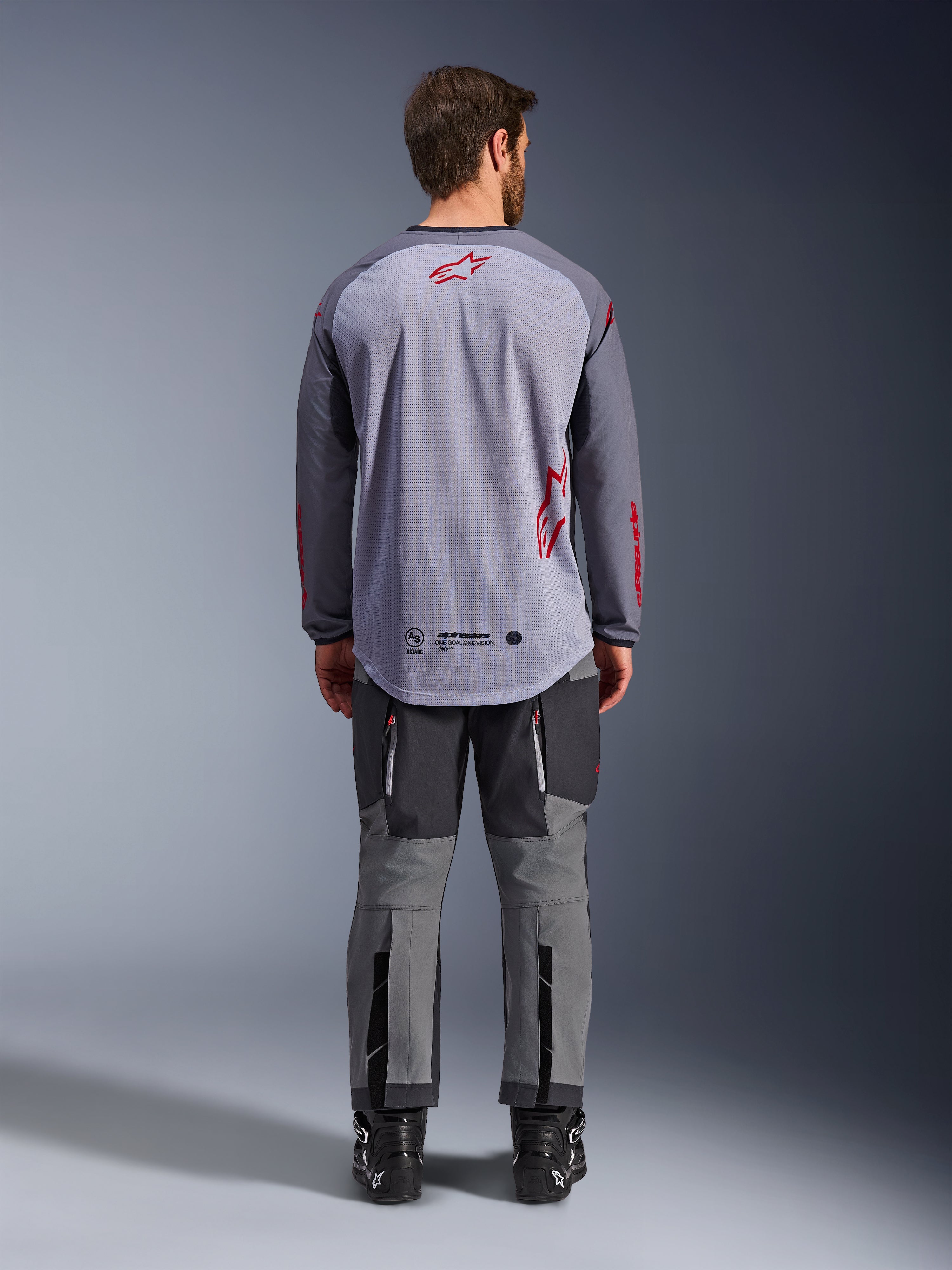 Mandlig model set bagfra iført Alpinestars Maxdura Dual Jersey, mellemgrå og mørkegrå adventure-trøje med røde logo-detaljer, mesh-rygkonstruktion, båret med matchende grå off-road bukser og sorte støvler