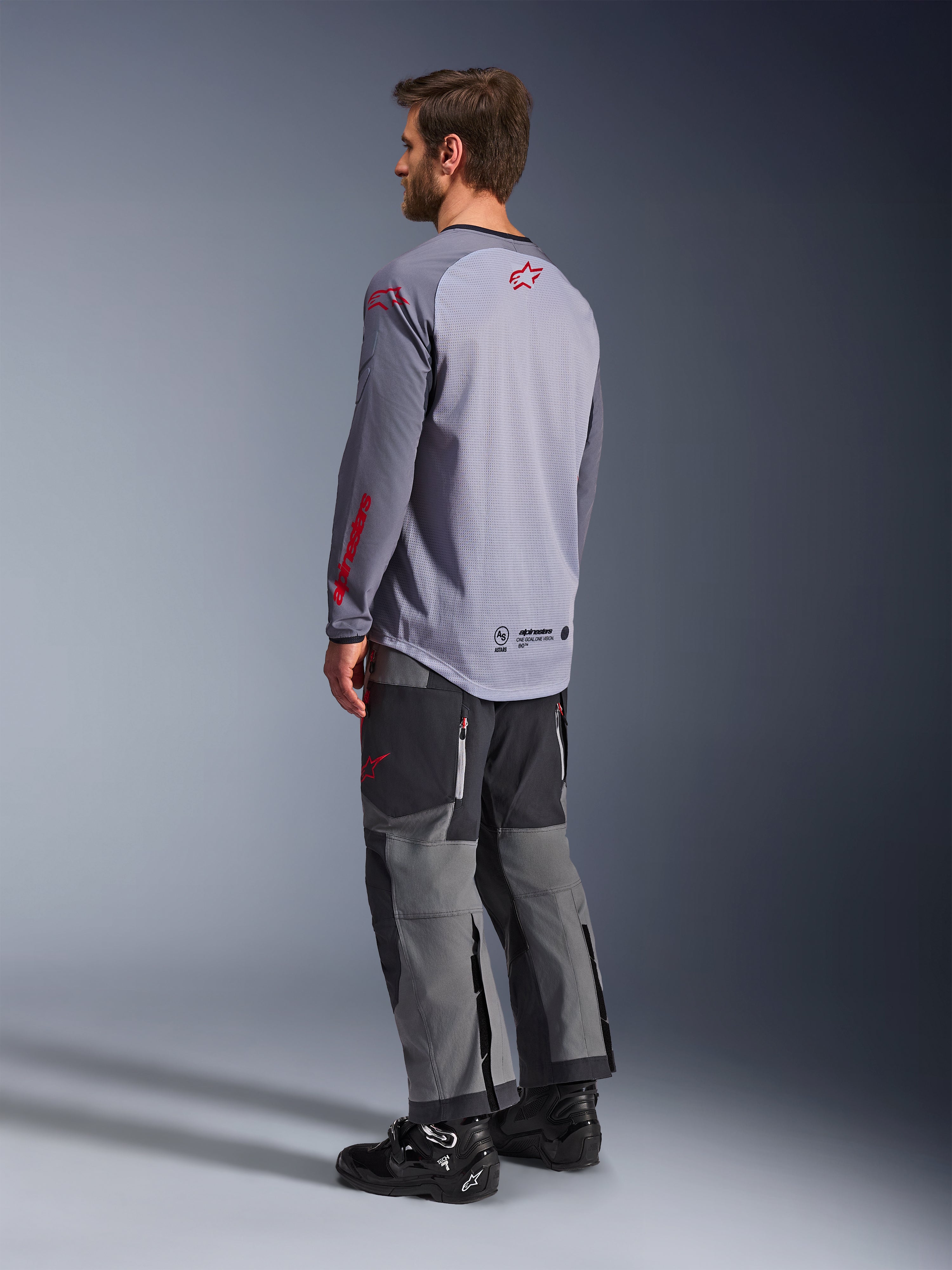 Mand iført Alpinestars Maxdura Dual Jersey, lysegrå med røde Alpinestars-logoer, set bagfra, viser mesh-overkrop og lange ærmer, kombineret med mørkegrå tekniske bukser og sorte motorcykelstøvler