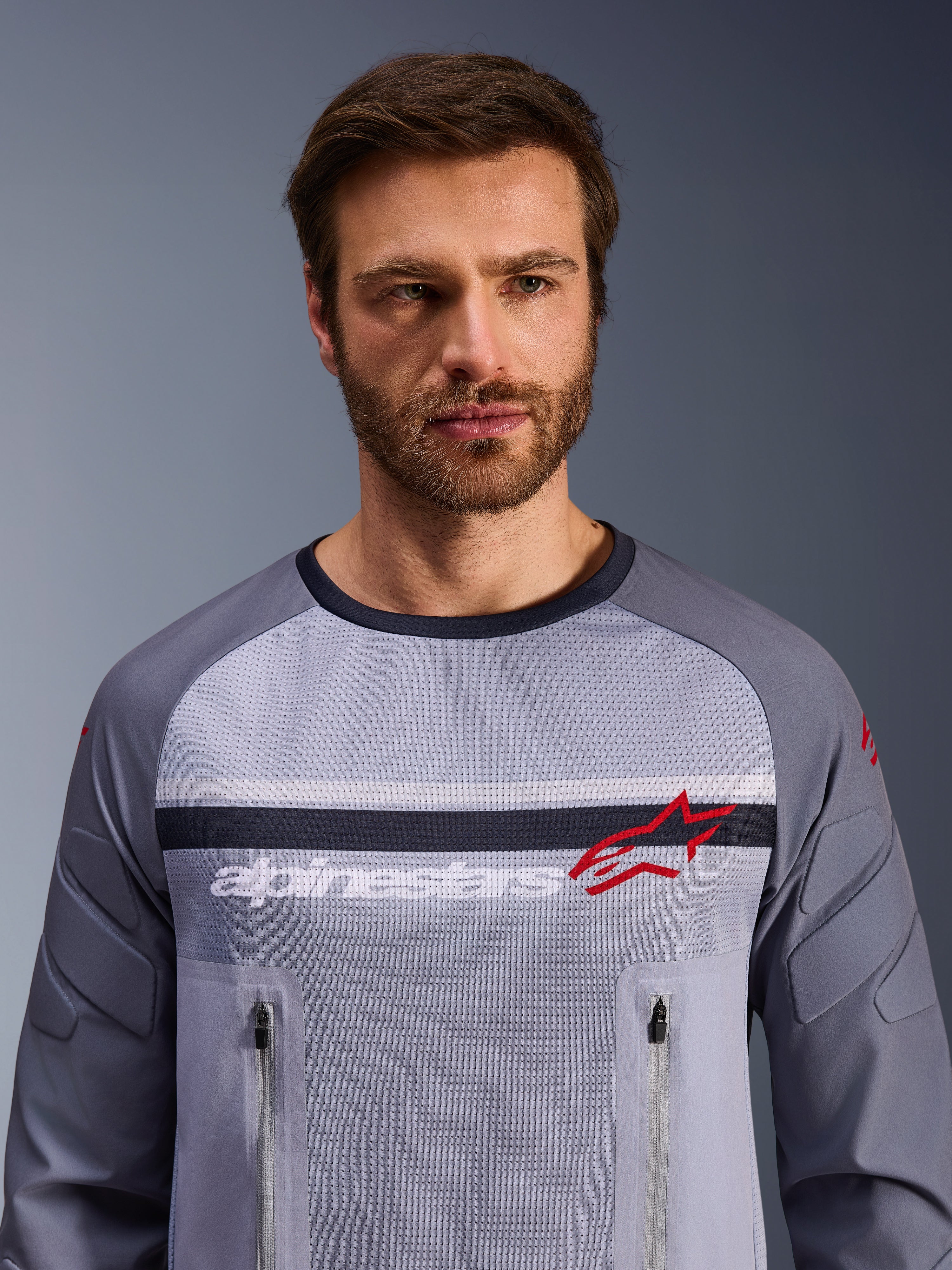 Alpinestars Maxdura Dual Jersey, mellemgrå og mørkegrå, båret af mandlig model, med mesh-torso til ventilation, dobbelte lynlåslommer på brystet, hvidt 'alpinestars'-branding og rødt stjernelogo, performance off-road adventure-udstyr