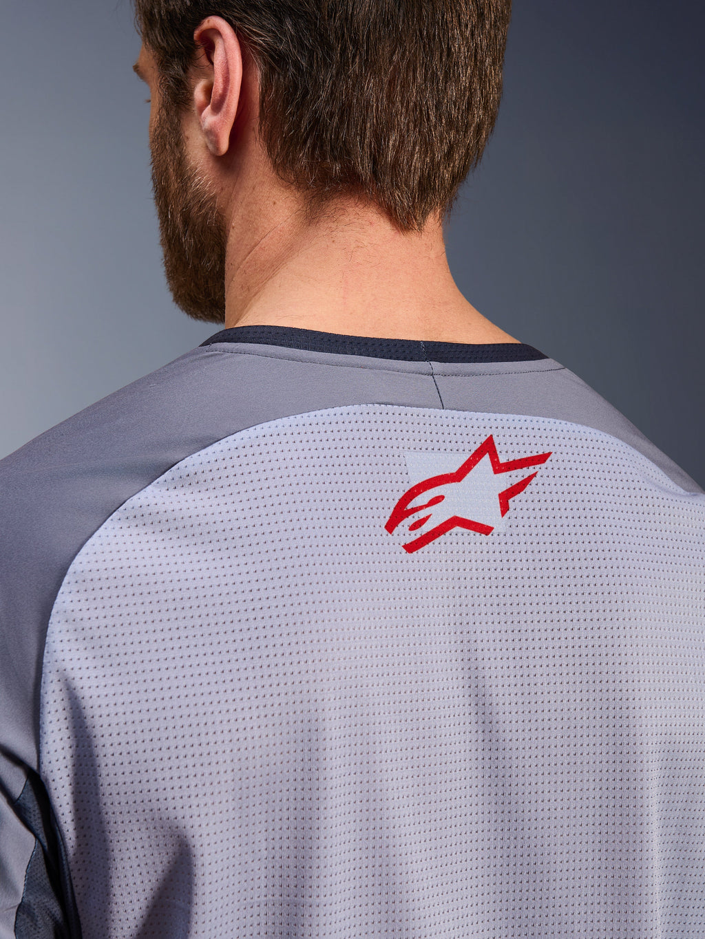 Alpinestars Maxdura Dual Jersey i mellemgrå og mørkegrå, nærbillede af den øverste del af ryggen og kraven, lille rødt Alpinestars-stjernelogo på åndbart lysegråt mesh-stof, mørkegrå kant ved halsen, designet til adventure- og off-road-kørsel