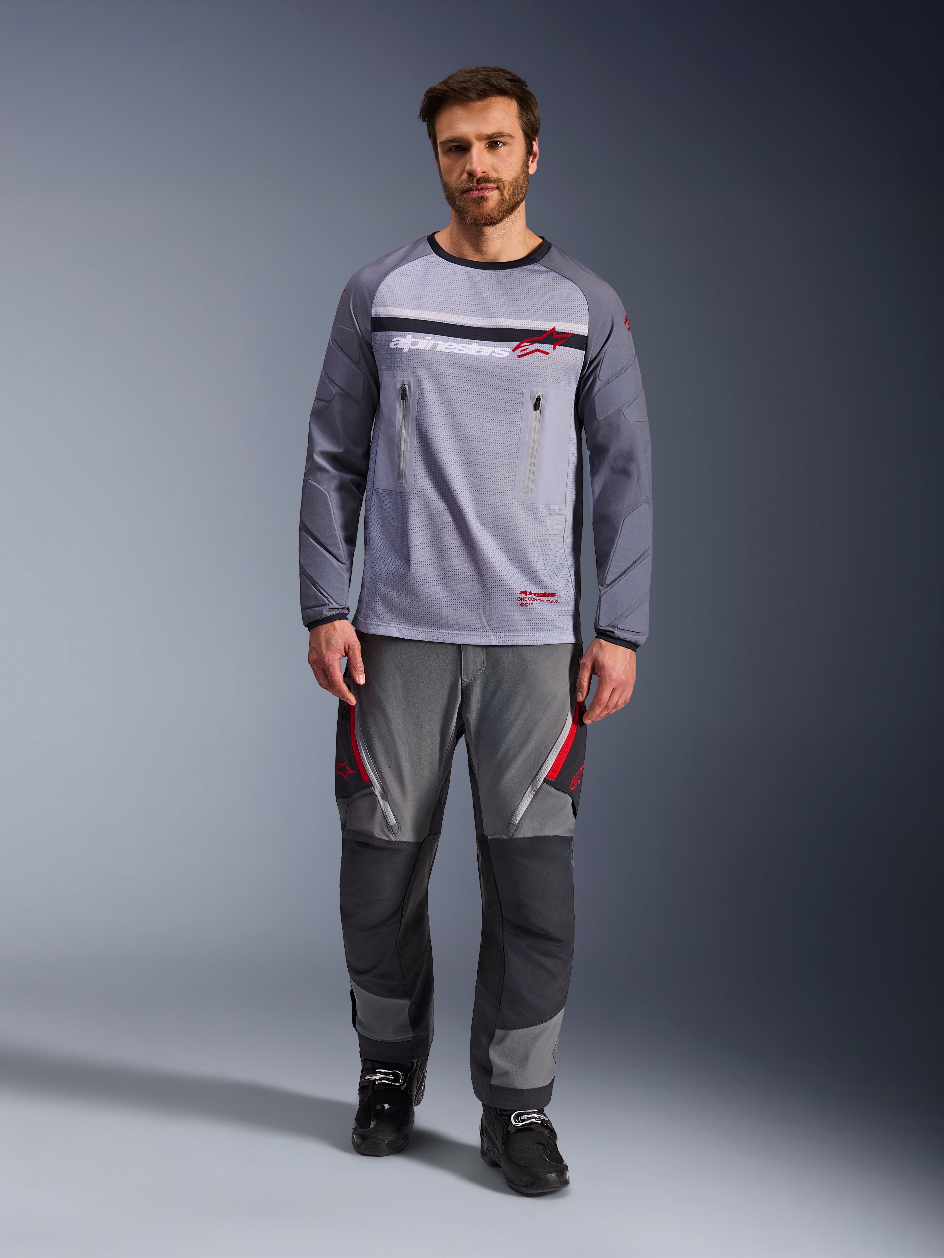 Alpinestars Maxdura Dual Jersey, mellemgrå og mørkegrå, adventure-motorcykeltrøje med mesh-overkrop og brystlommer med lynlås, slidstærke ærmer med integreret polstring, båret med matchende grå performance-bukser med røde detaljer, teknisk off-road-køreudstyr