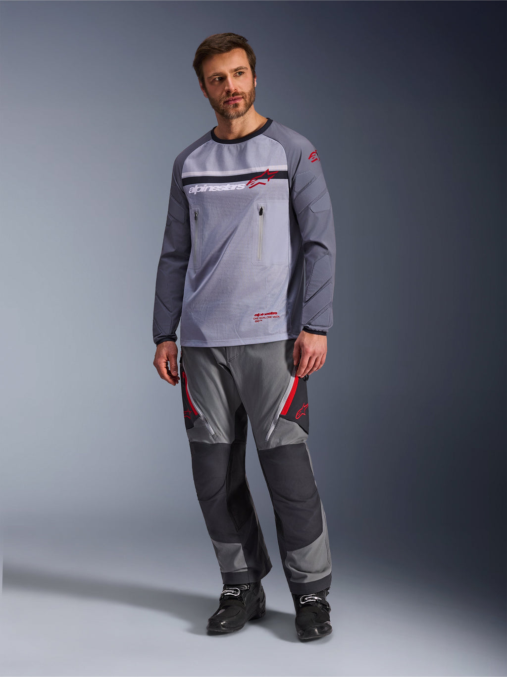 En mand iført Alpinestars Maxdura Dual Jersey i mellemgrå og mørkegrå, stylet med matchende grå og sorte adventure-bukser og sorte støvler, med en åndbar mesh-overkrop, brystlommer med lynlås og røde logo-detaljer.