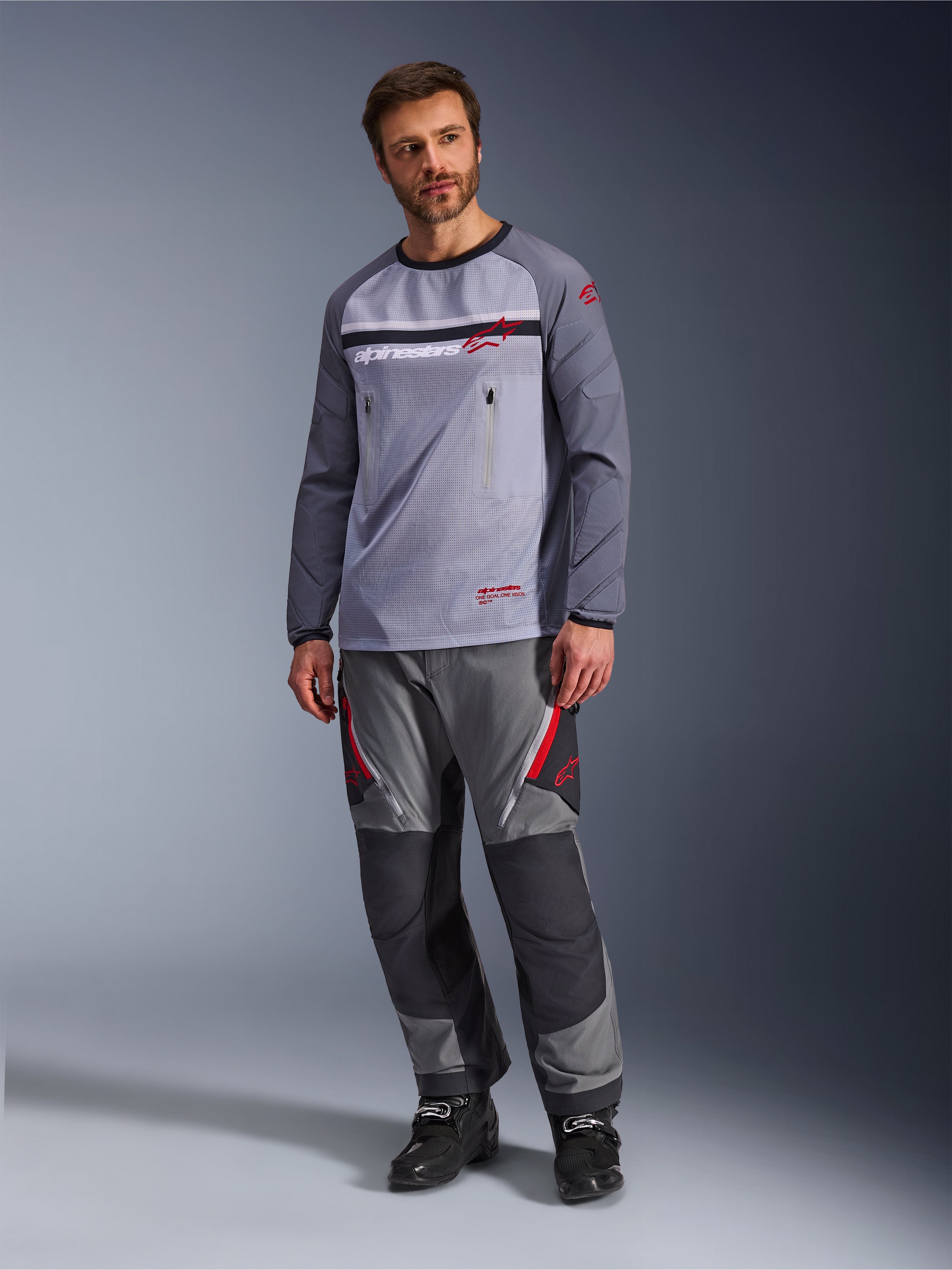 En mand iført Alpinestars Maxdura Dual Jersey i mellemgrå og mørkegrå, stylet med matchende grå og sorte adventure-bukser og sorte støvler, med en åndbar mesh-overkrop, brystlommer med lynlås og røde logo-detaljer.