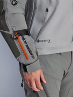Alpinestars Nazca 3L GORE-TEX PRO jakke, mørkegrå og mellemgrå adventure-jakke, nærbillede af ærme med sort branding og orange lynlåsdetalje, GORE-TEX PRO-logo på torso, holdbar tekstilkonstruktion med forstærket albuepanel