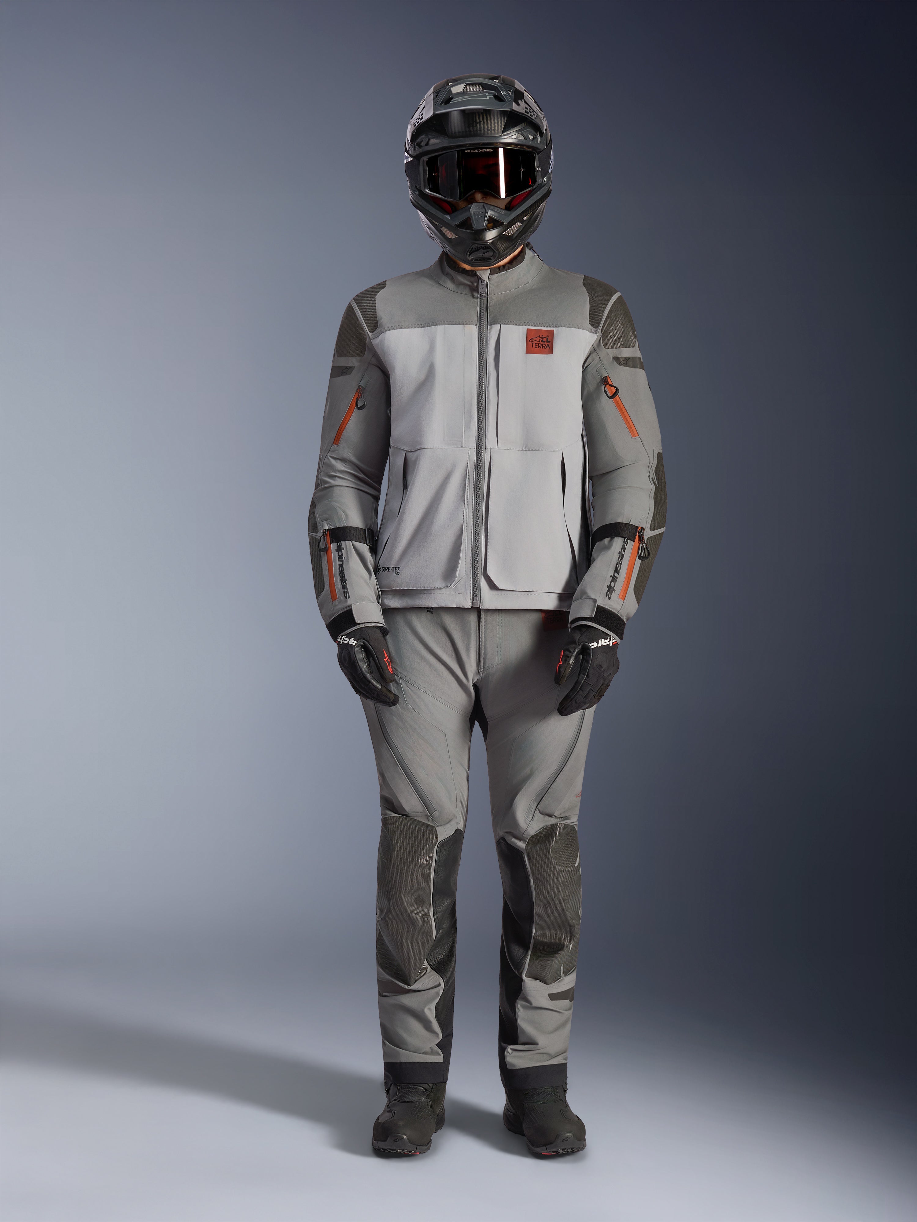 Alpinestars Nazca 3L GORE-TEX PRO Jakke, adventure motorcykeljakke, mørkegrå og mellemgrå med orange detaljer, med Superfabric-forstærkninger og ventilationslynlåse foran, båret med matchende bukser og sort hjelm, teknisk terrænkørselsudstyr