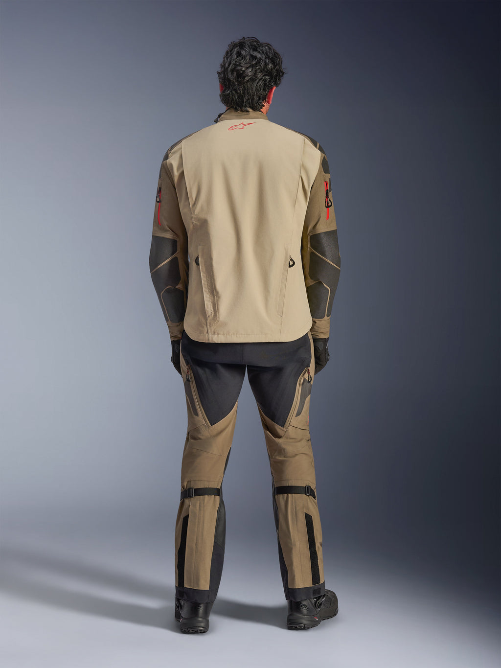 Alpinestars Nazca 3L GORE-TEX PRO Jakke i Walnut Rock Brown, set bagfra på kører, viser Superfabric-forstærkninger på albuerne og bageste ventilationslynlåse, båret med matchende adventure-bukser, sorte støvler og handsker, robust teknisk køreudstyr til ekstrem adventure.